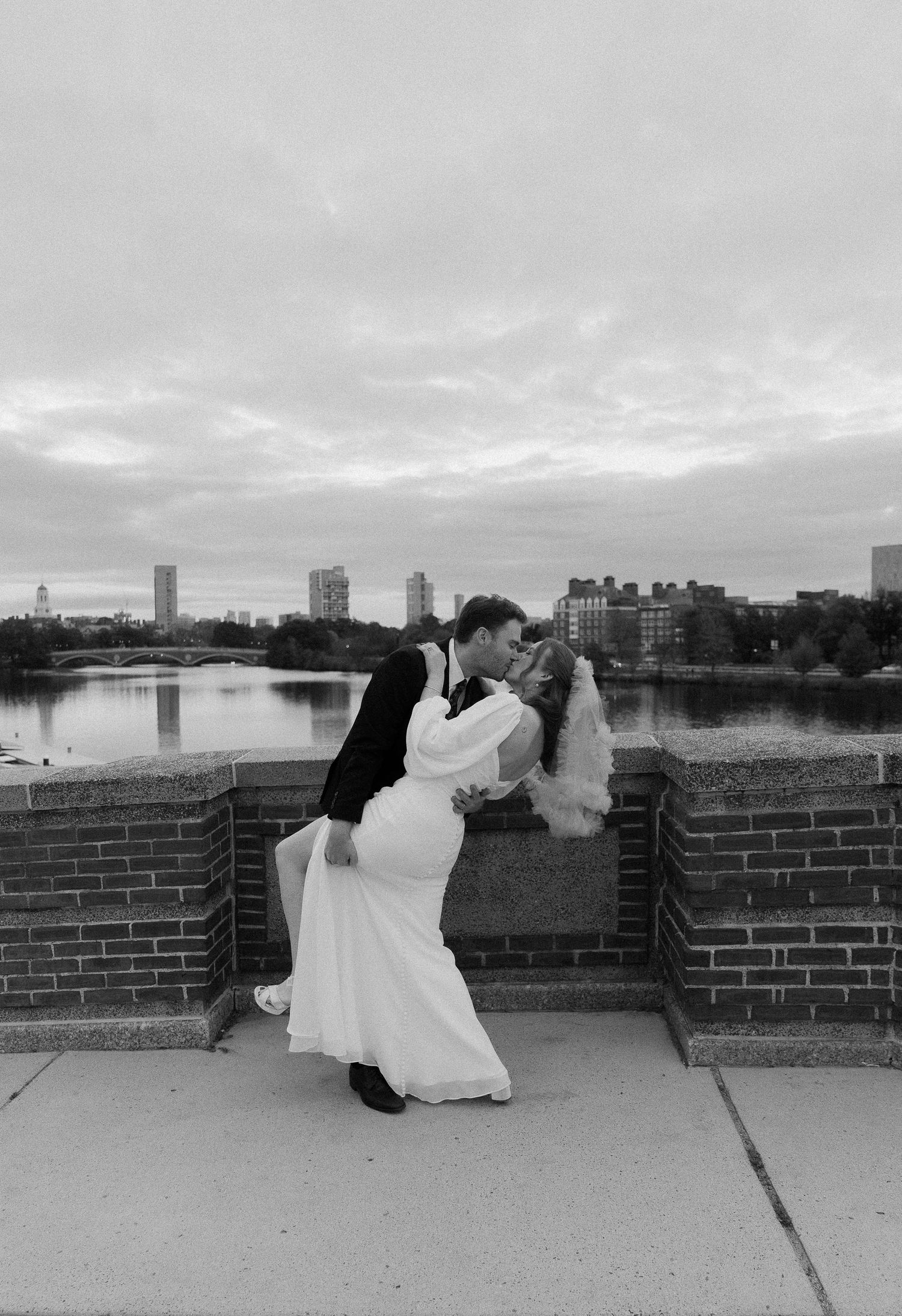 Black and white photo of a bride and groom sharing a romantic dip kiss on a bridge with a city skyline backdrop.