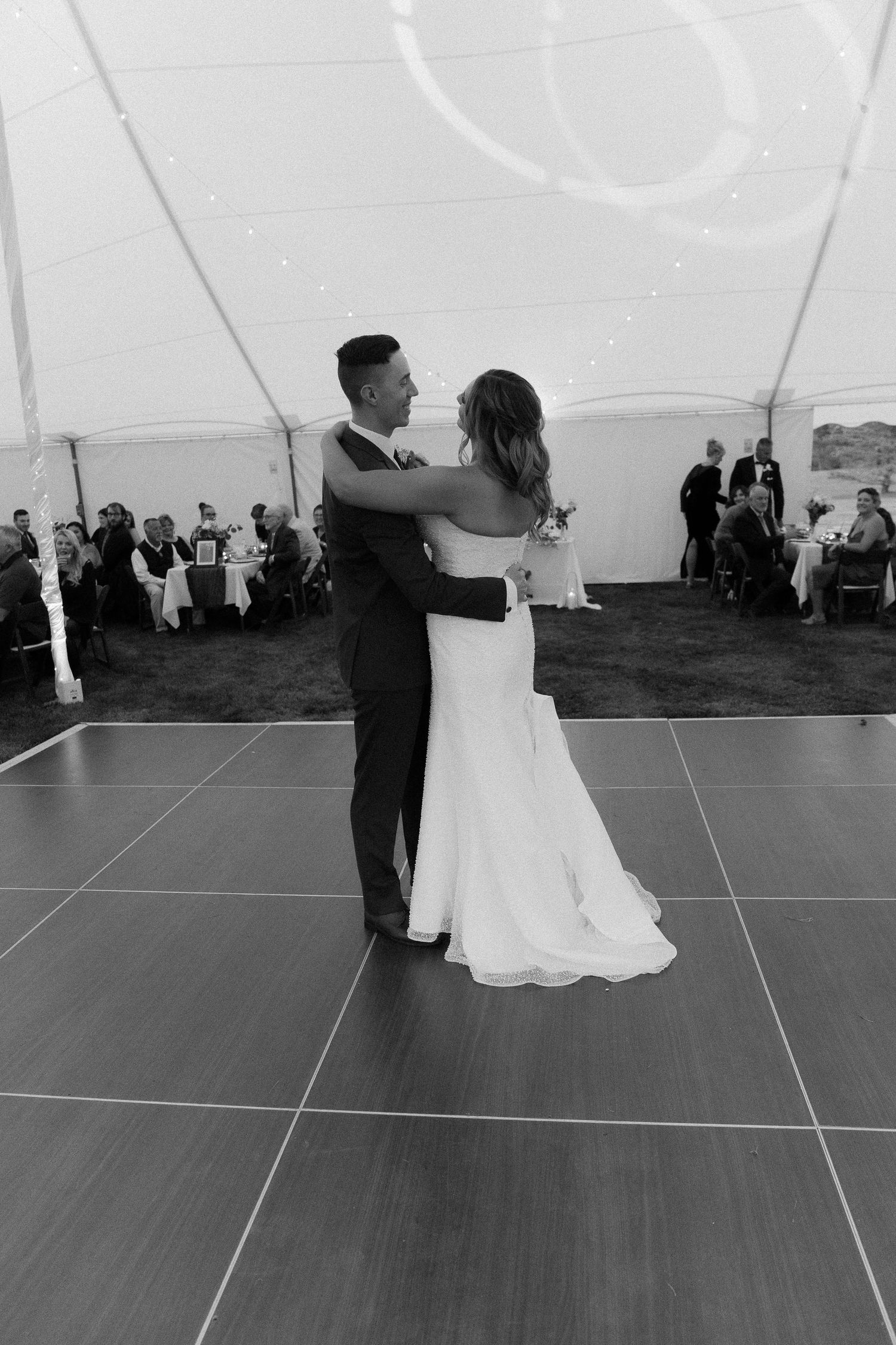 A couple dances together on a dark wooden floor under a white tent at their wedding reception.