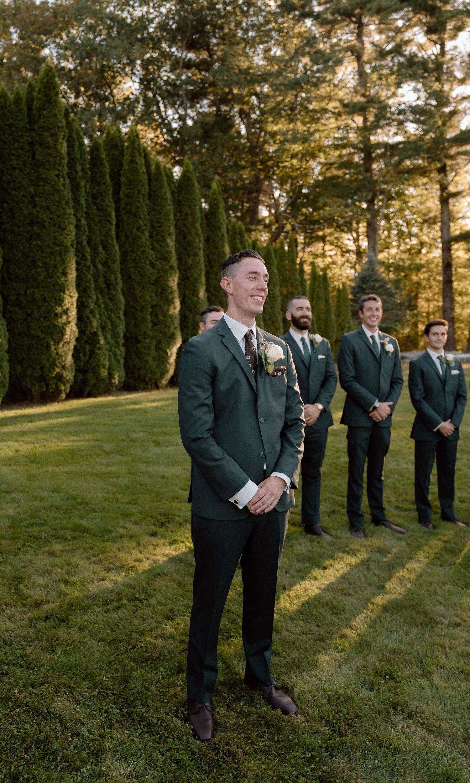 Groomsmen share laughs while standing in formation during outdoor sunset ceremony.