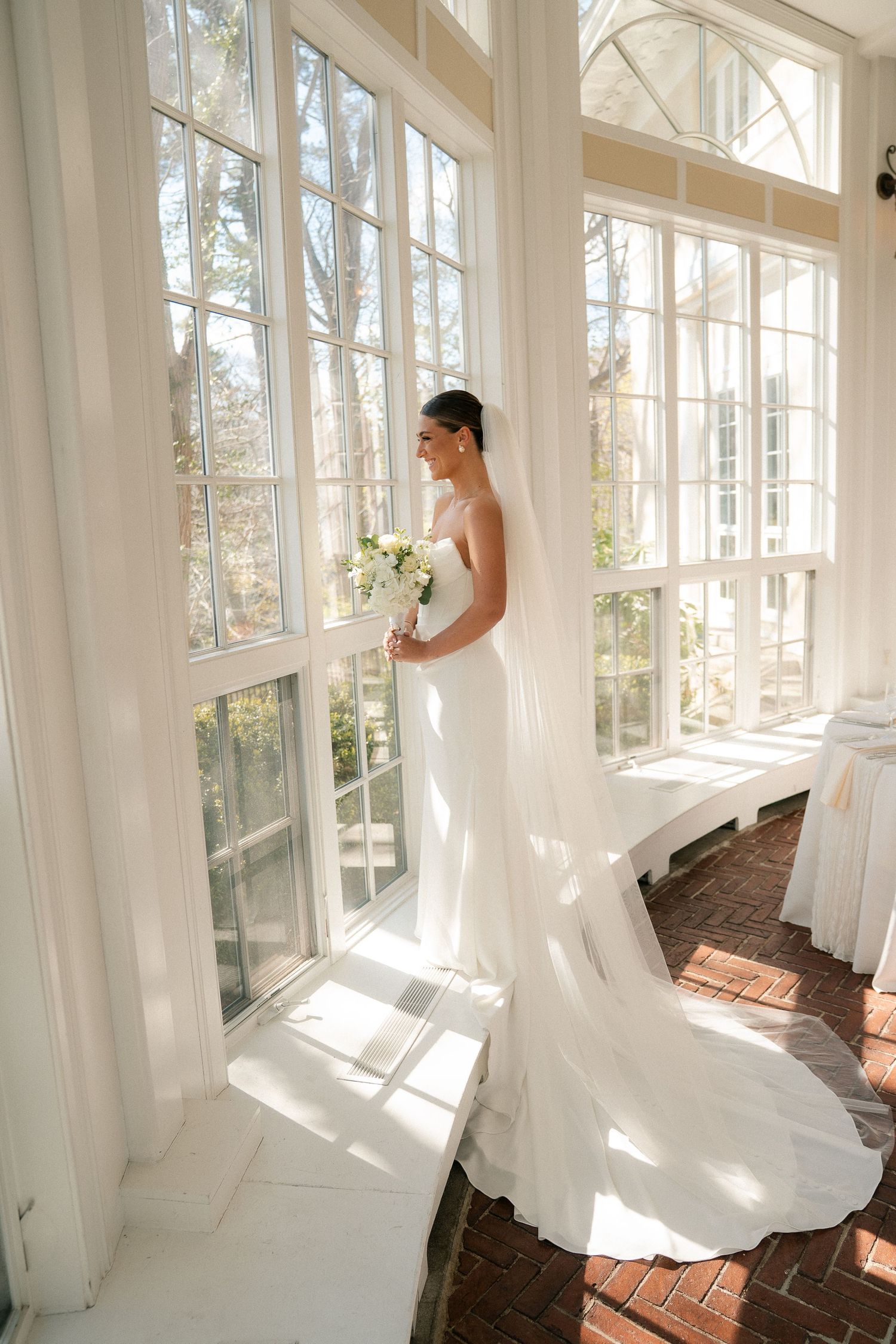 Bride in white gown with train holds bouquet by sunlit conservatory windows on her wedding day.