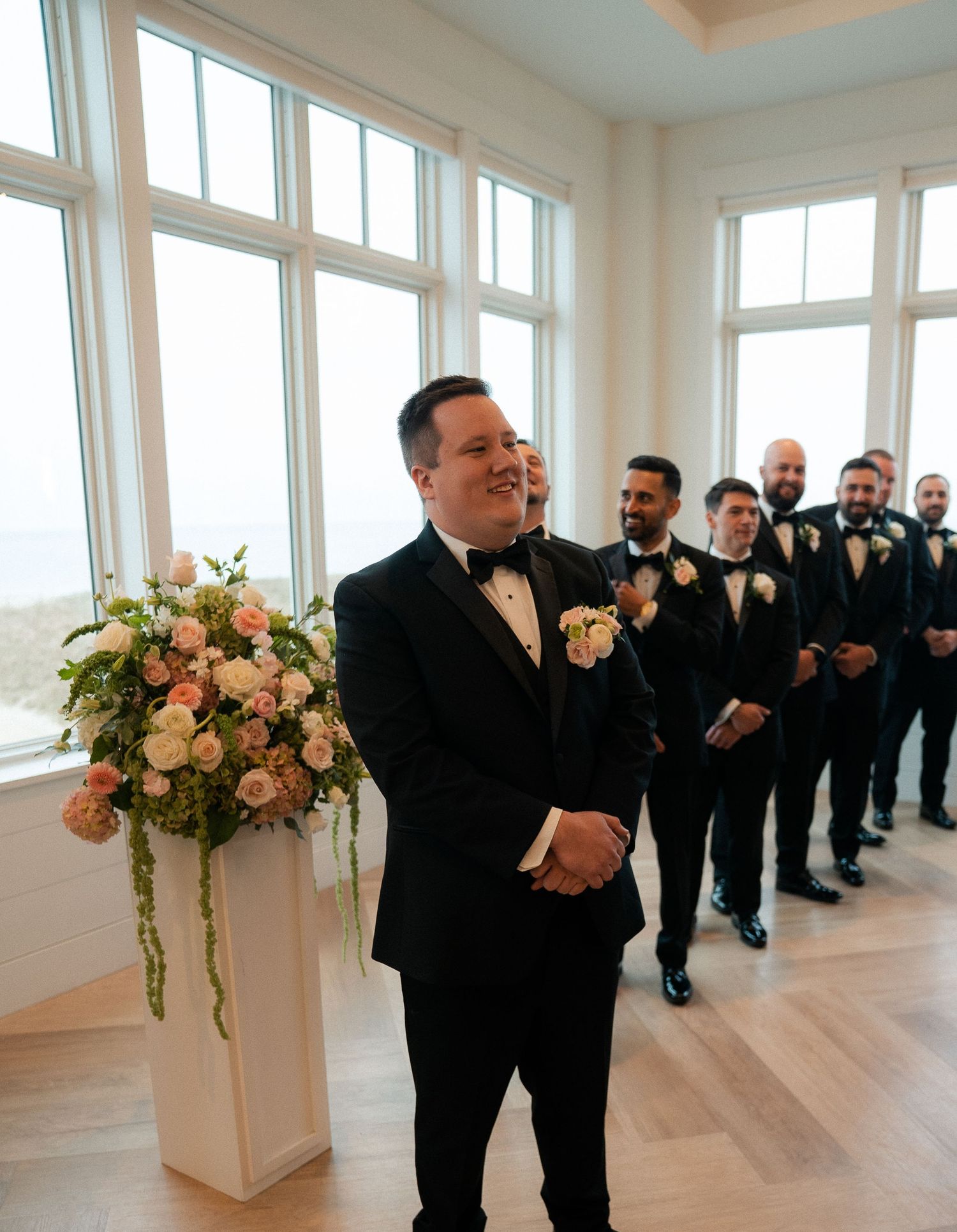 Groom in black tuxedo smiling at altar with groomsmen lined up beside floral arrangement at wedding ceremony.