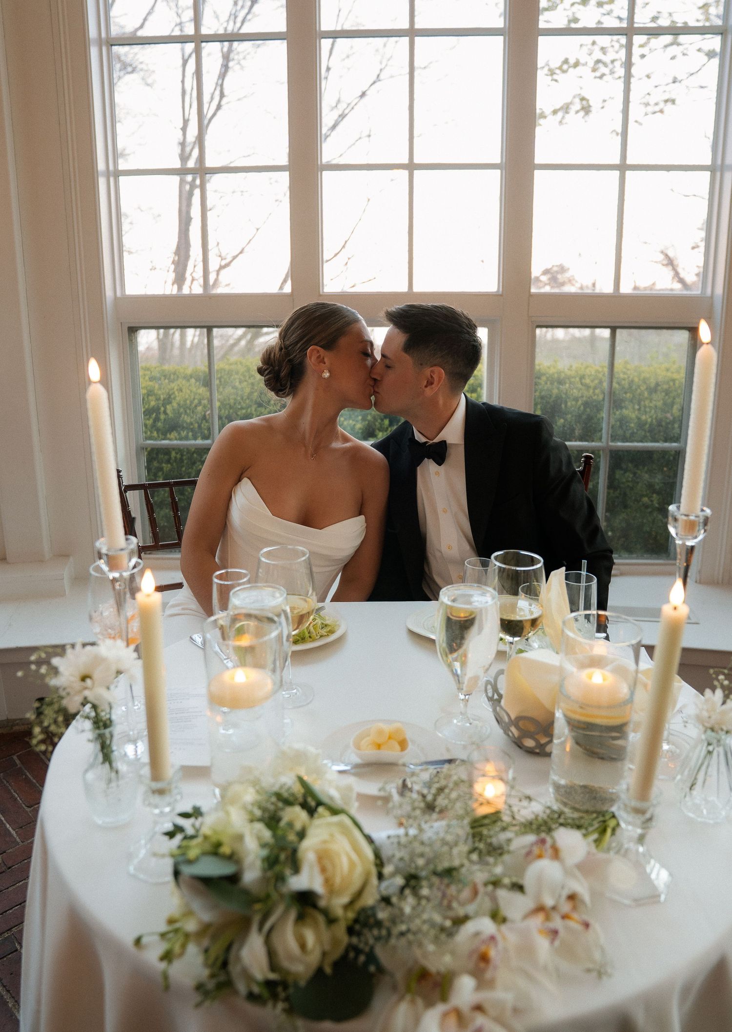 Bride and groom sharing a kiss at candlelit sweetheart table with white roses at their wedding reception.