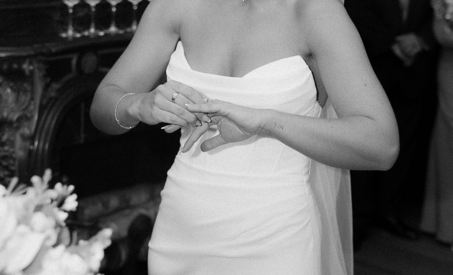 Black and white photo of a bride in a strapless white gown placing a wedding ring on her finger.