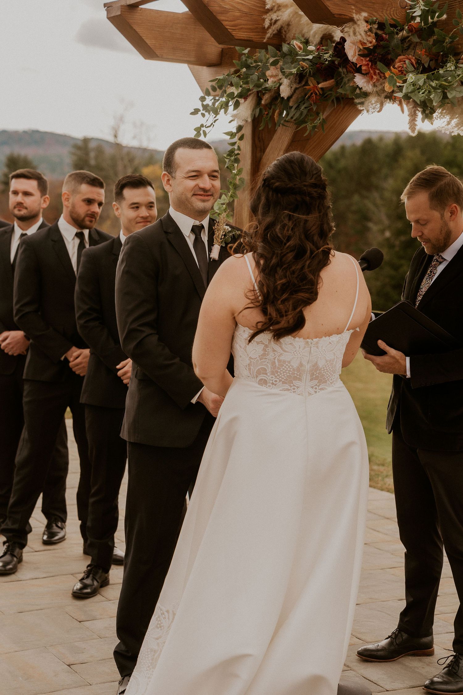 Wedding ceremony taking place under a wooden arbor decorated with floral arrangements on a stone patio.