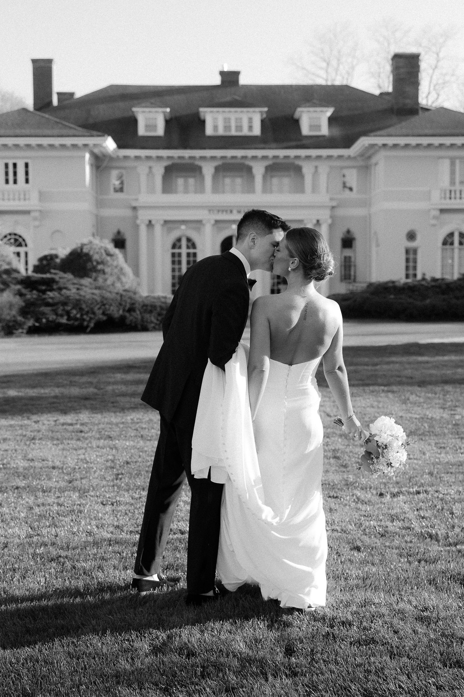 Bride and groom kiss on lawn before grand estate mansion in classic black and white wedding photo.