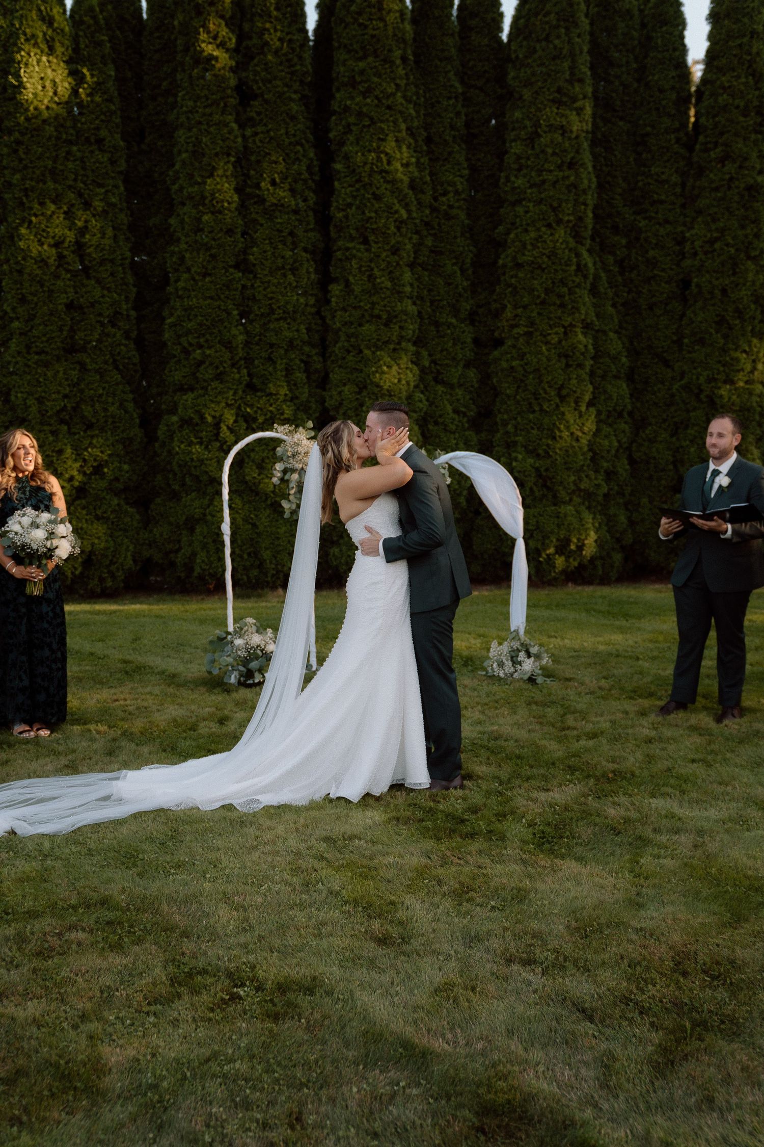A wedding ceremony takes place outdoors with white decorations and tall evergreen trees in the background.