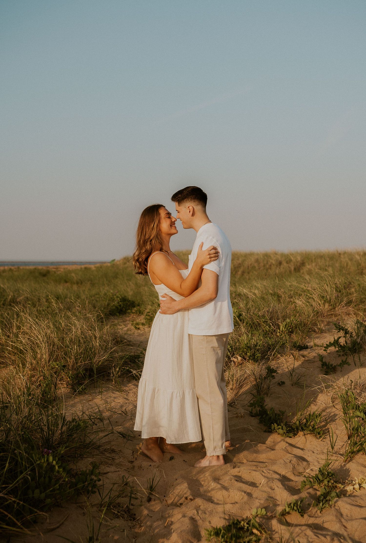Sunset engagement photos of a couple in light-colored outfits standing on sandy terrain with prairie grass.