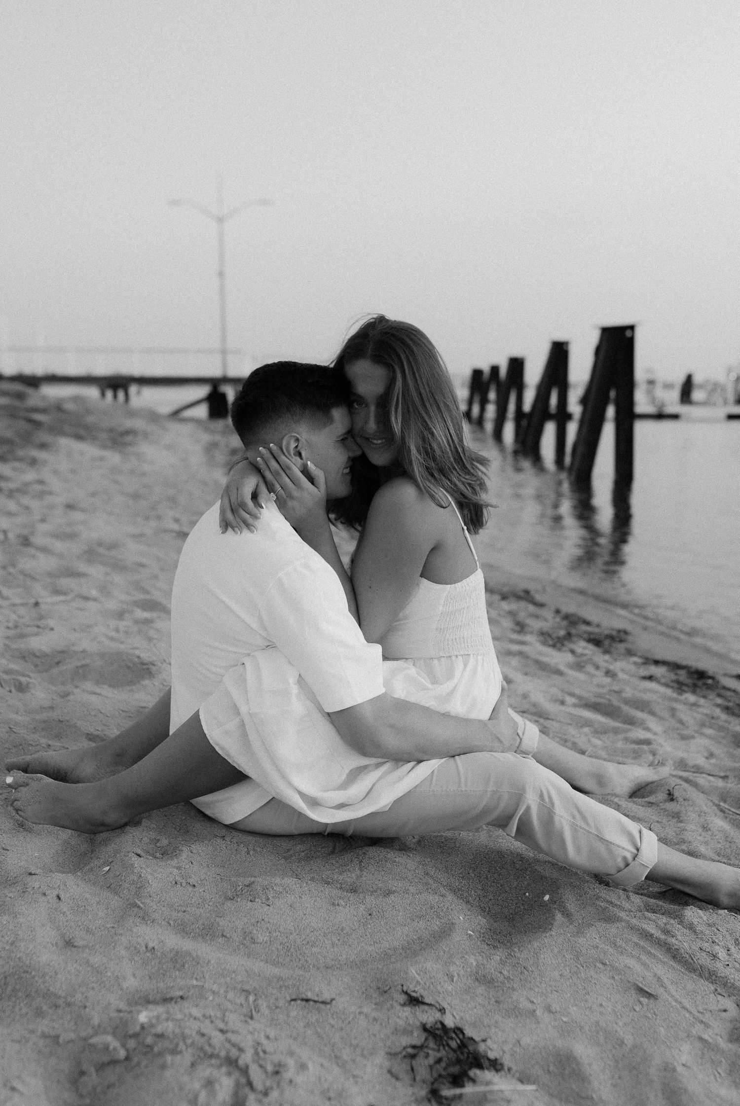 Black and white portrait of couple embracing on beach near wooden pier posts.