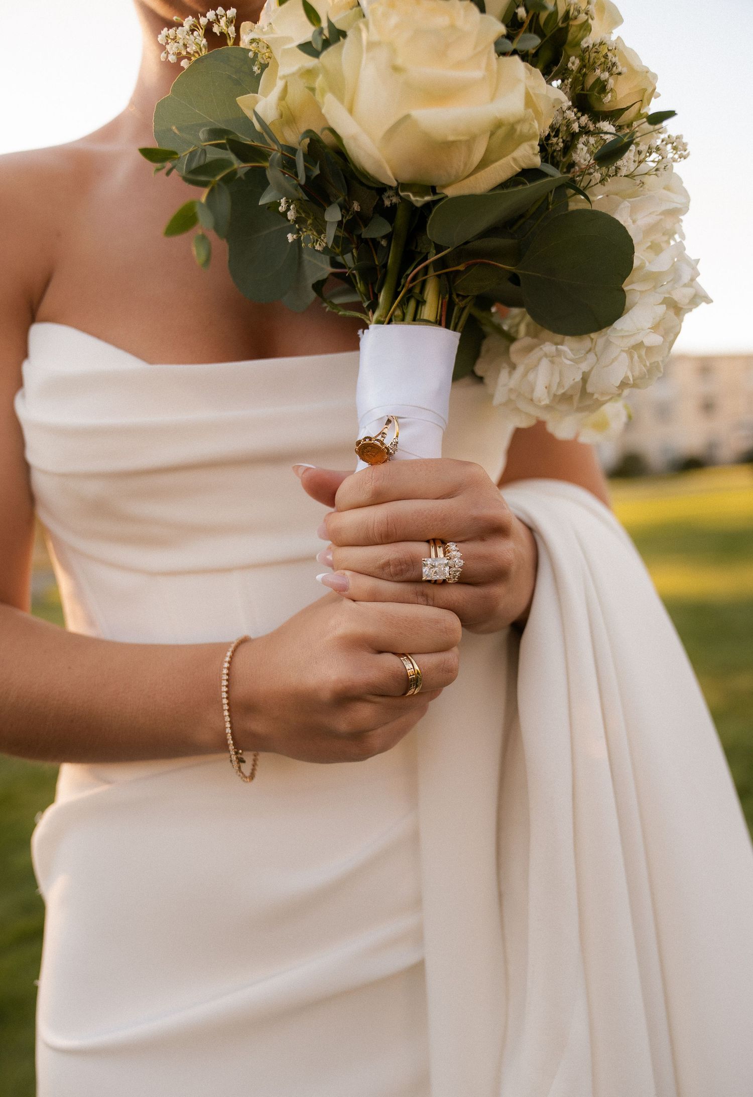 Bride in strapless white gown holding ivory rose bouquet, showcasing diamond engagement ring and gold bracelet.