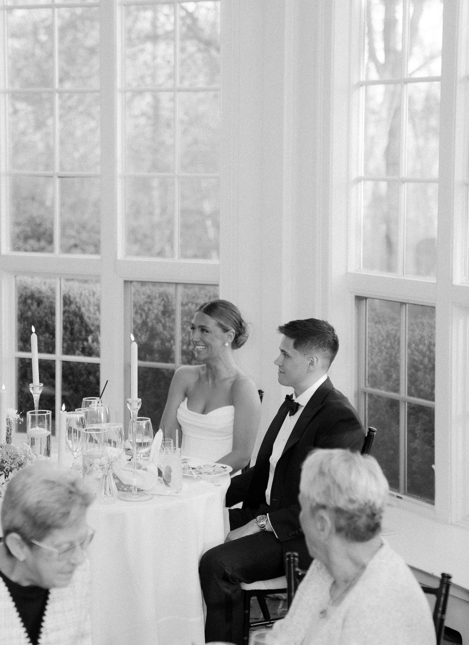 Bride and groom sit together at sweetheart table during wedding reception near large windows in black and white photo.