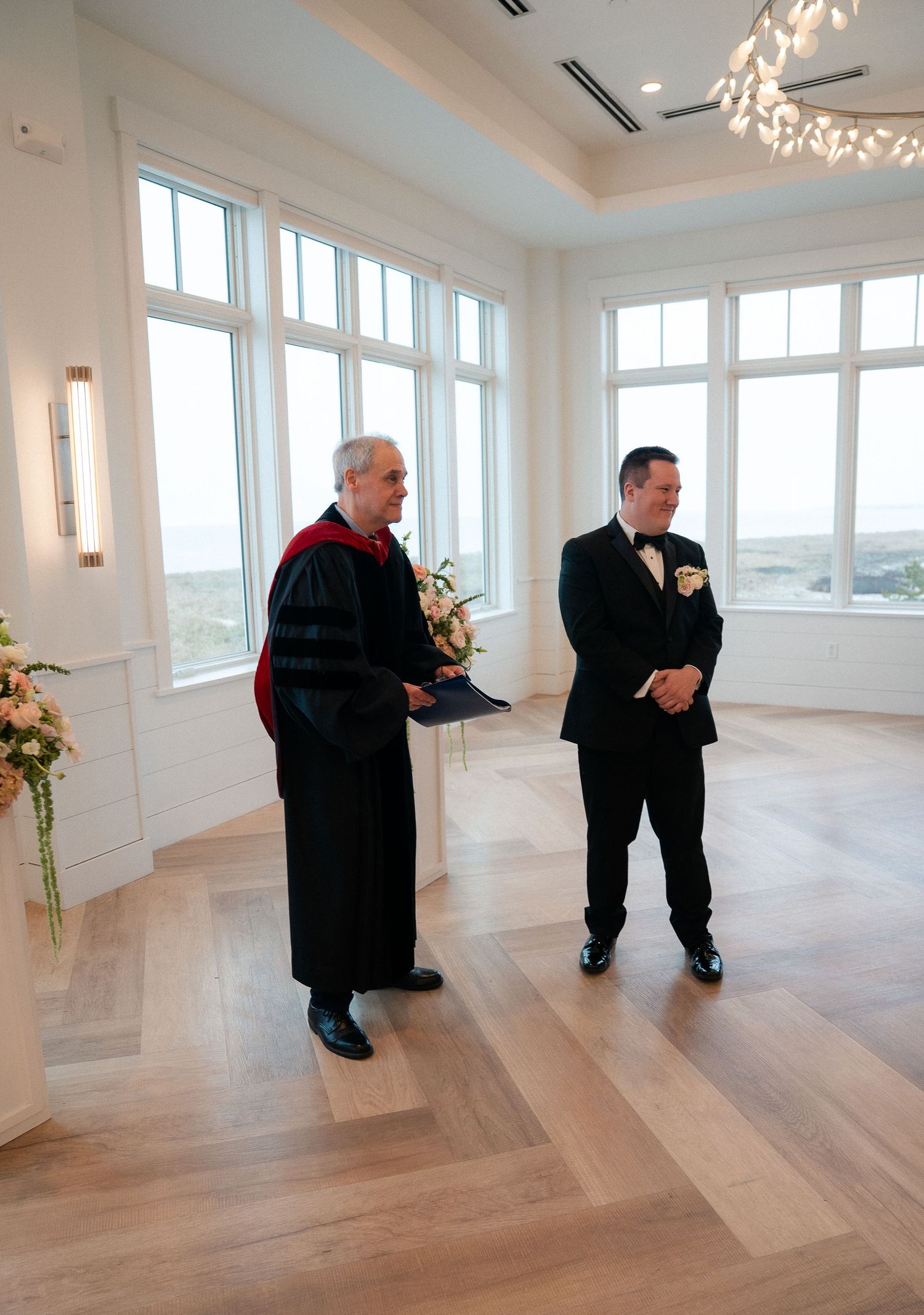 Groom in black tuxedo stands with officiant in academic robes during indoor wedding ceremony with ocean views.