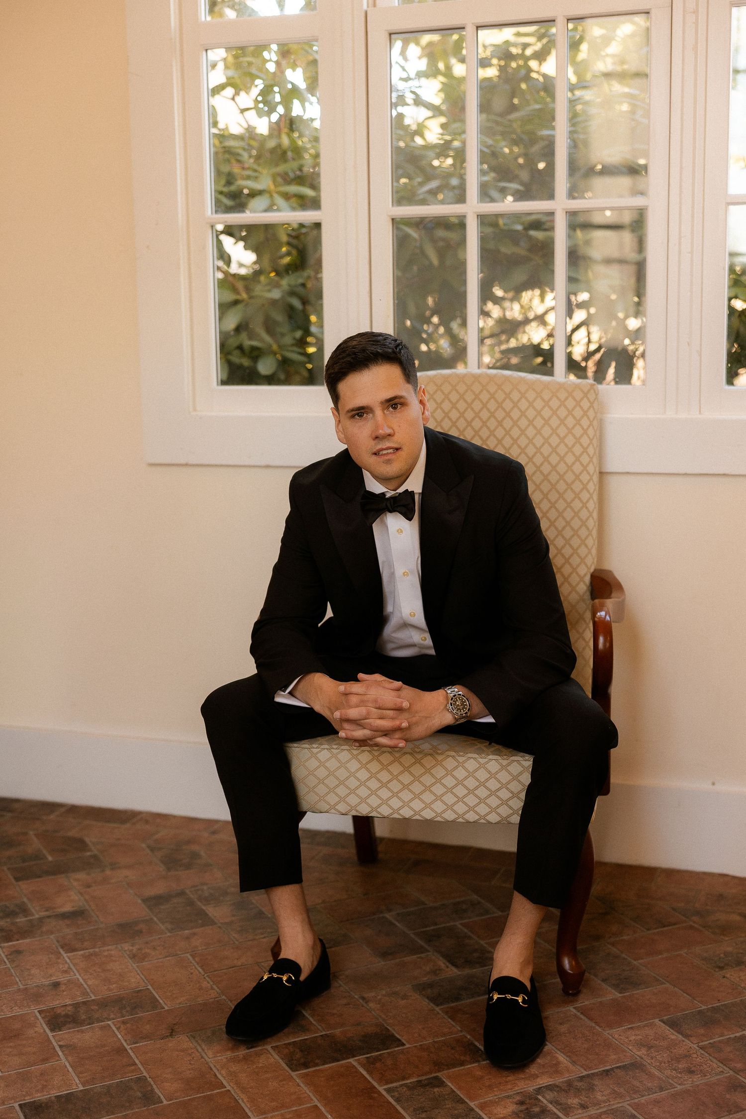A well-dressed man in a black tuxedo and bow tie sits confidently in an elegant chair by a bright window.