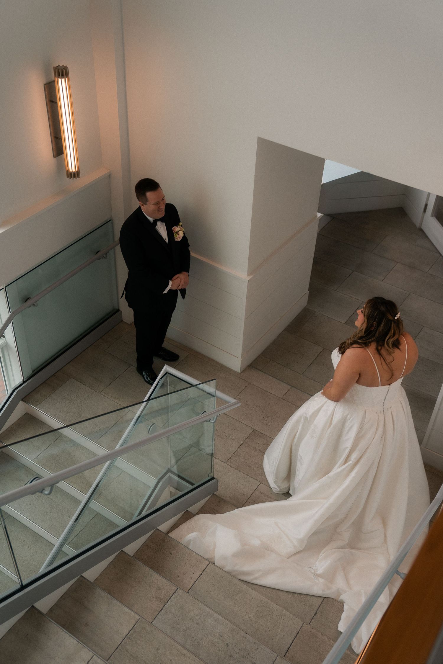 Bride in white gown and groom in black tuxedo share a moment on modern staircase with glass railings.