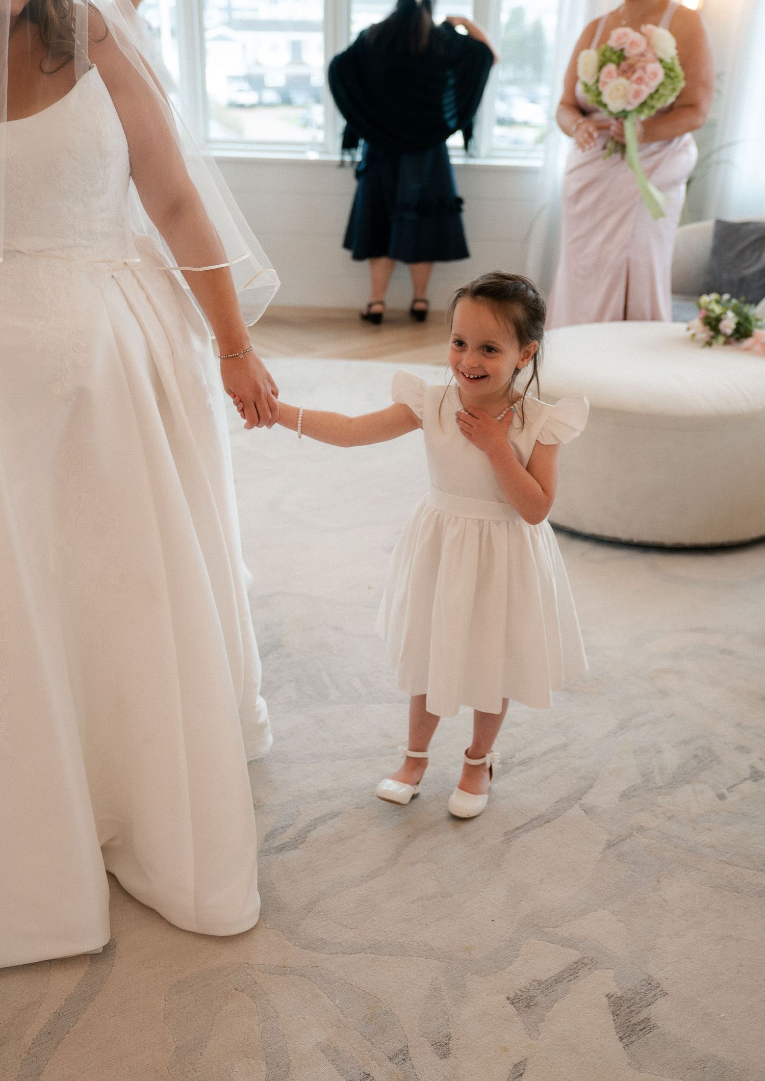 A young flower girl in a white dress holds the bride's hand, smiling joyfully during wedding preparations indoors.