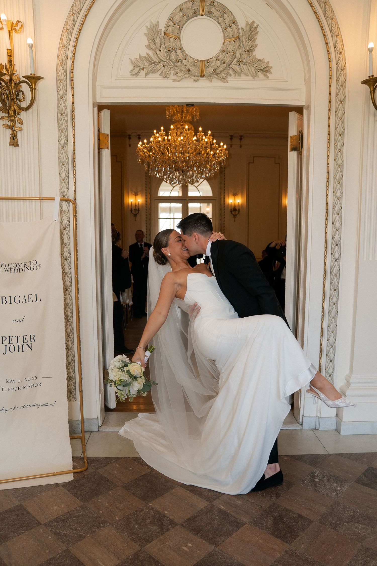Bride and groom share a romantic kiss in an elegant ballroom doorway, with the groom lifting his bride in her white gown.