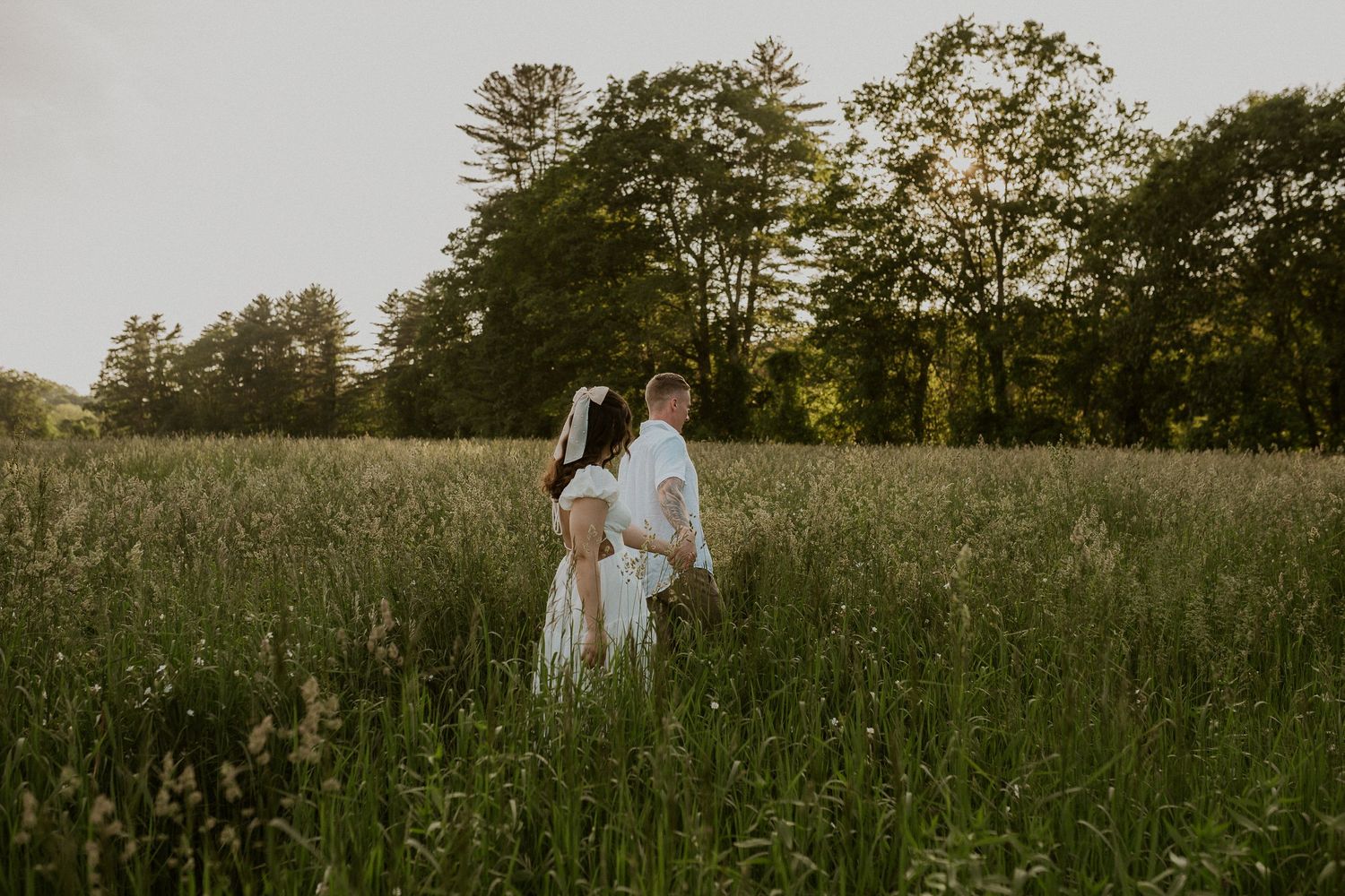 A couple stands together in a tall grass meadow during golden hour.