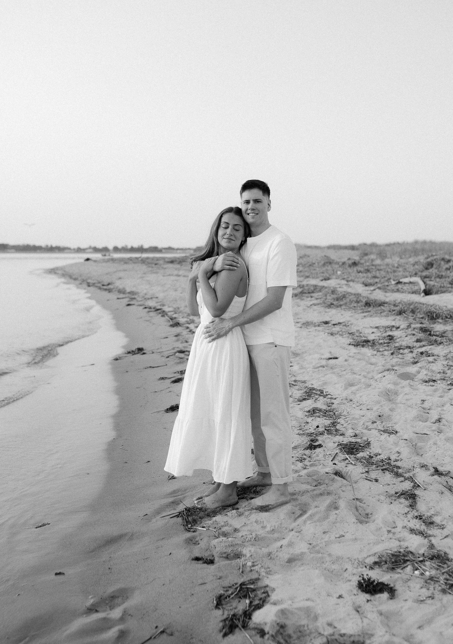 Black and white photo of a couple embracing on a beach during a sunset photoshoot.