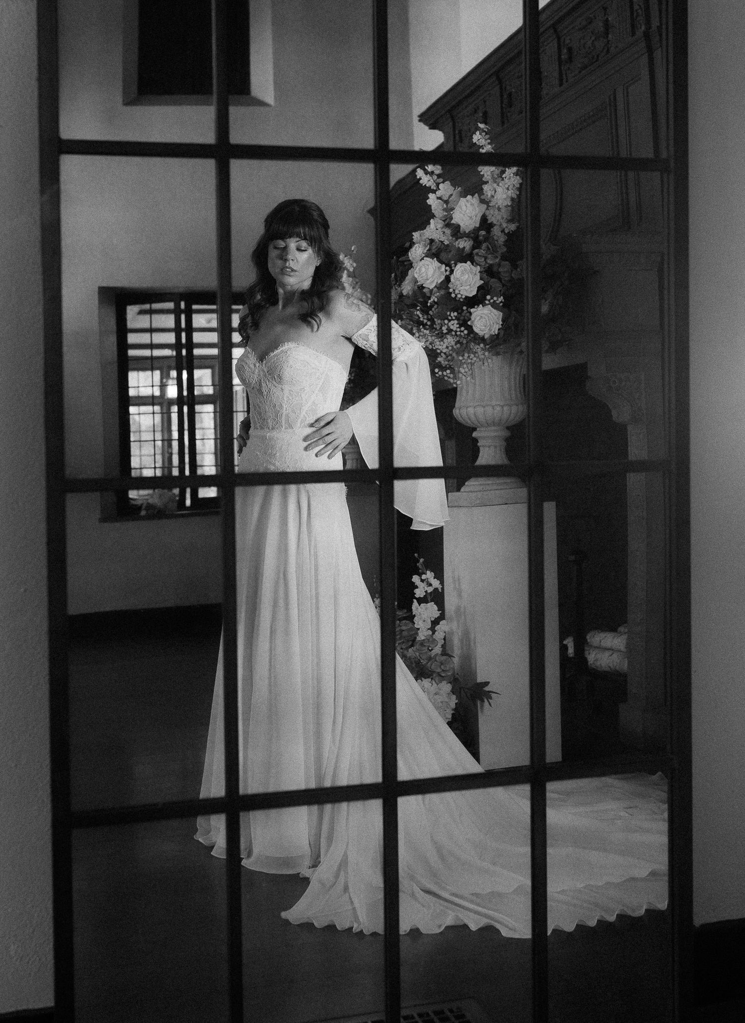 Black and white portrait of bride in flowing gown viewed through window panes.
