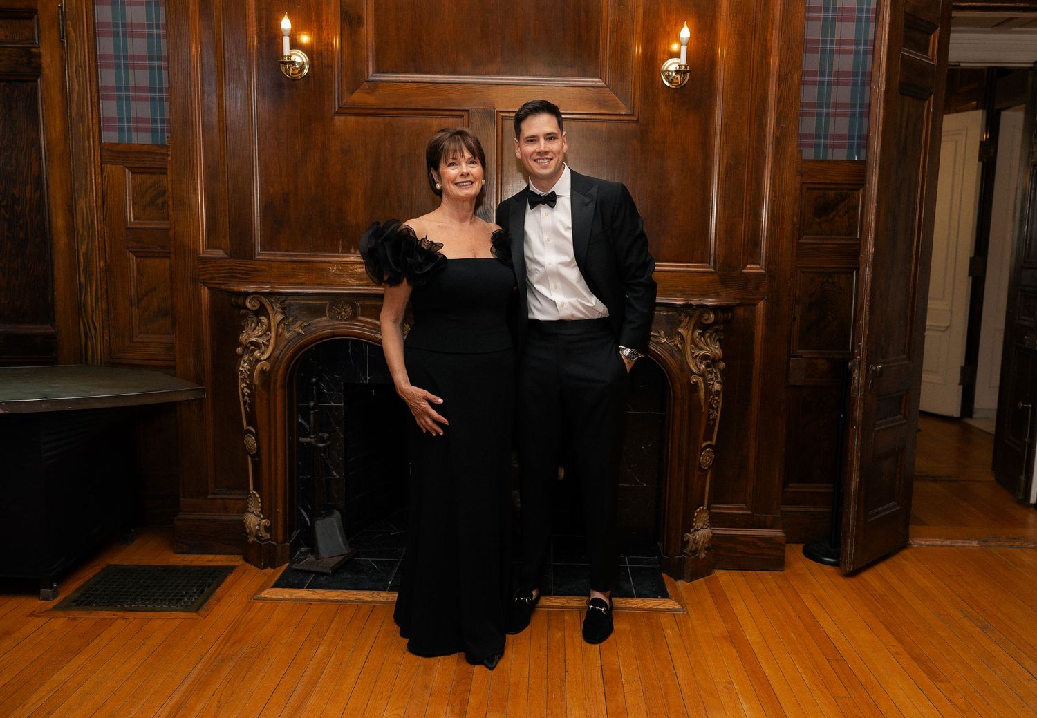 Elegant couple in formal black tie attire posing in front of ornate wood-paneled fireplace at upscale event.