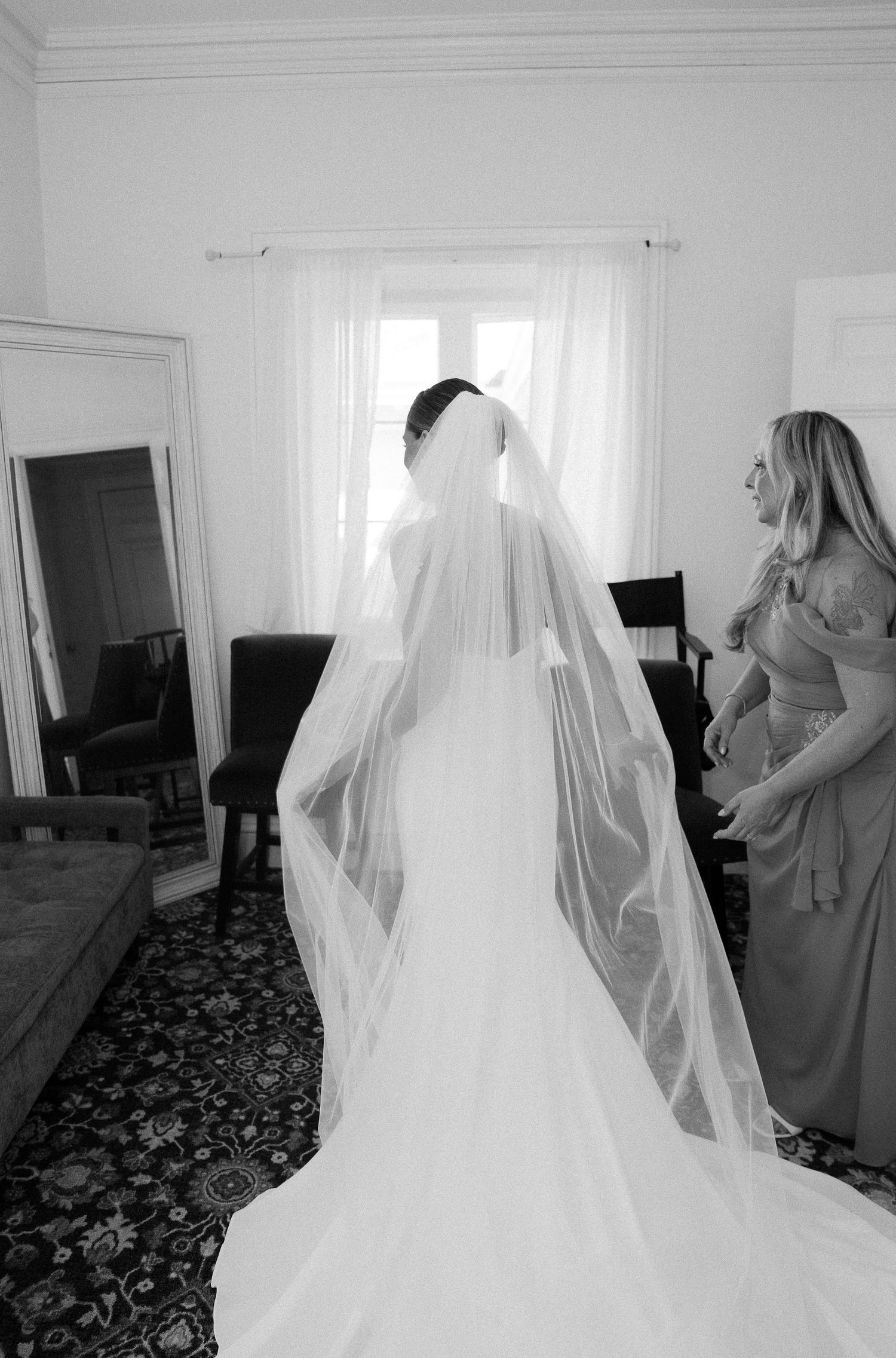 Bride in wedding gown with long veil standing by window with bridesmaid, black and white photo.