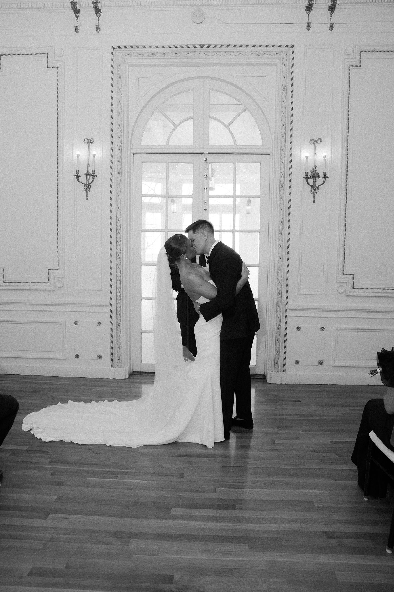 Bride and groom share a romantic kiss in a stunning white ballroom in black and white photo.