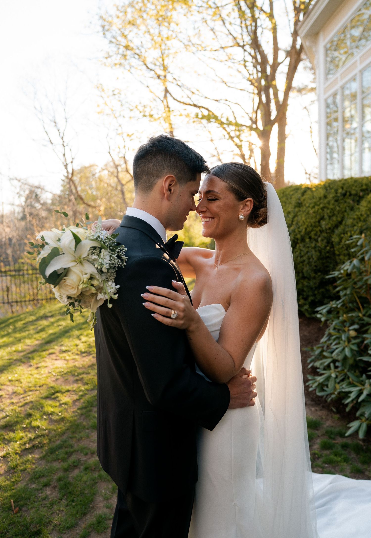 A bride and groom share a romantic moment outdoors, with golden sunlight and lush greenery in the background.