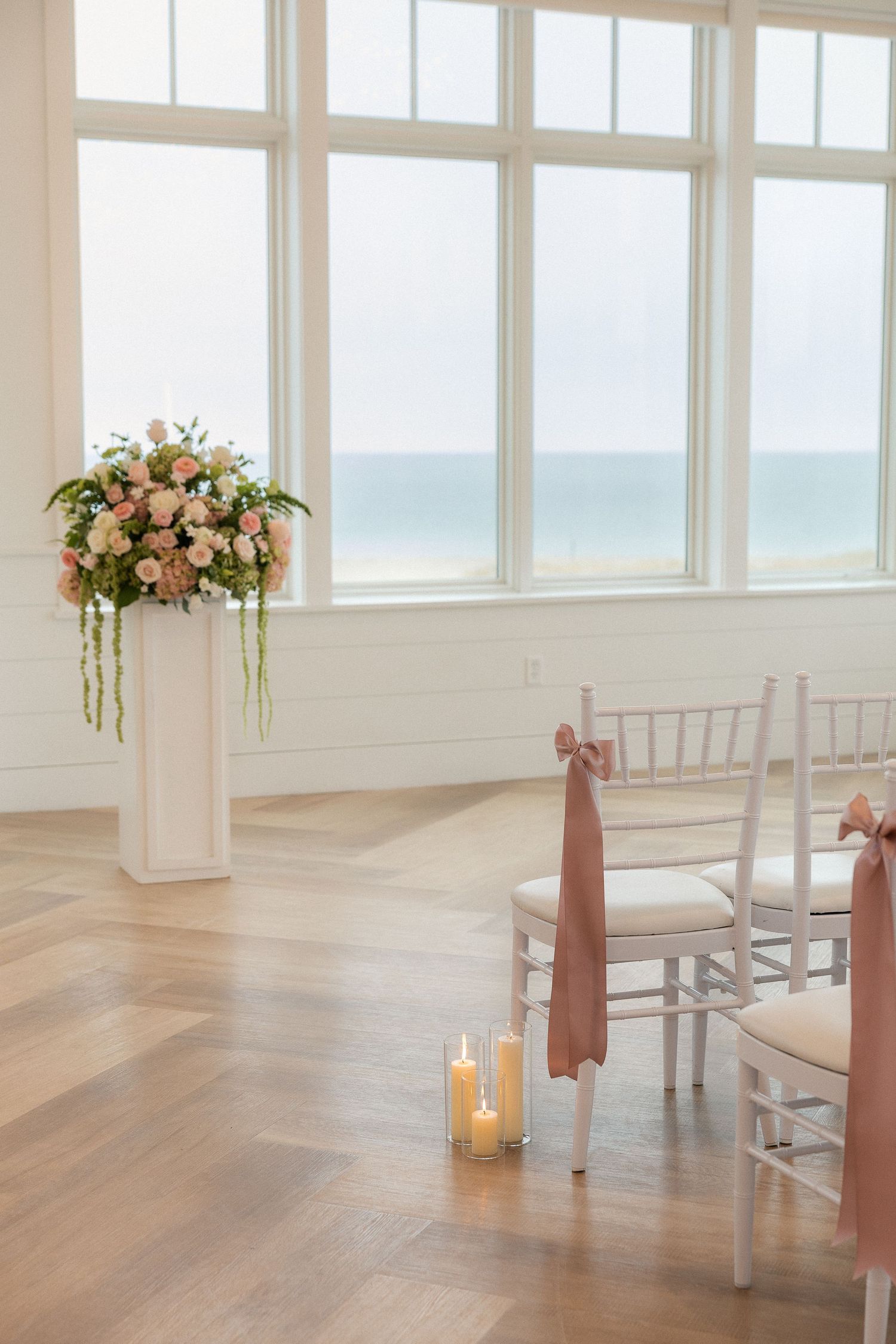 Elegant beachfront wedding venue with floral arrangements, candles, and chiavari chairs with blush ribbon bows.
