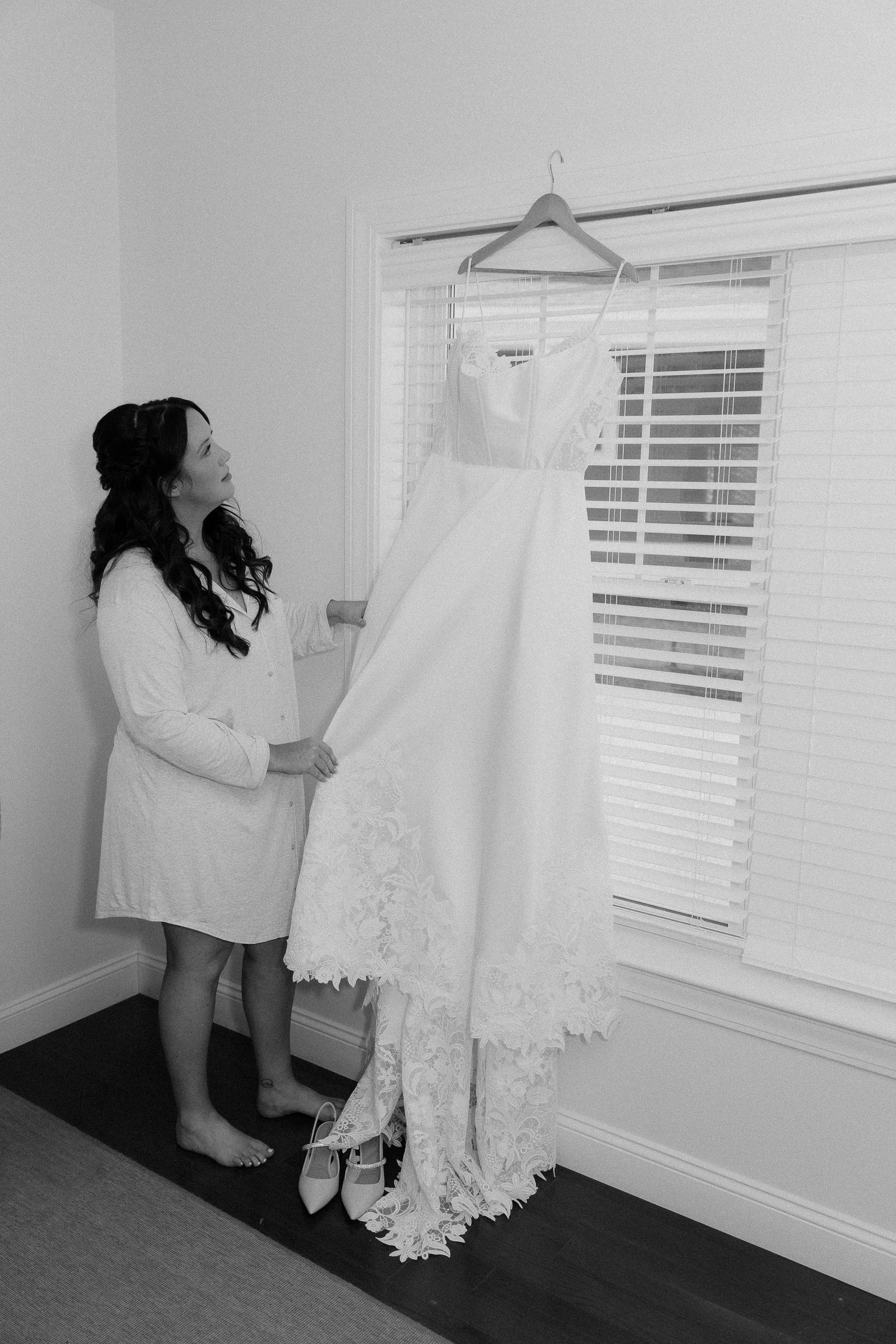 A white lace wedding dress hangs by a window while someone admires it in a robe.