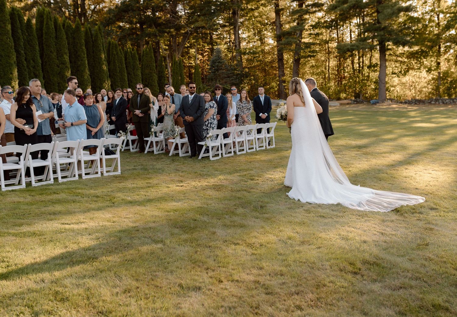 Outdoor wedding ceremony setup with white chairs and guests on a spacious lawn at golden hour.