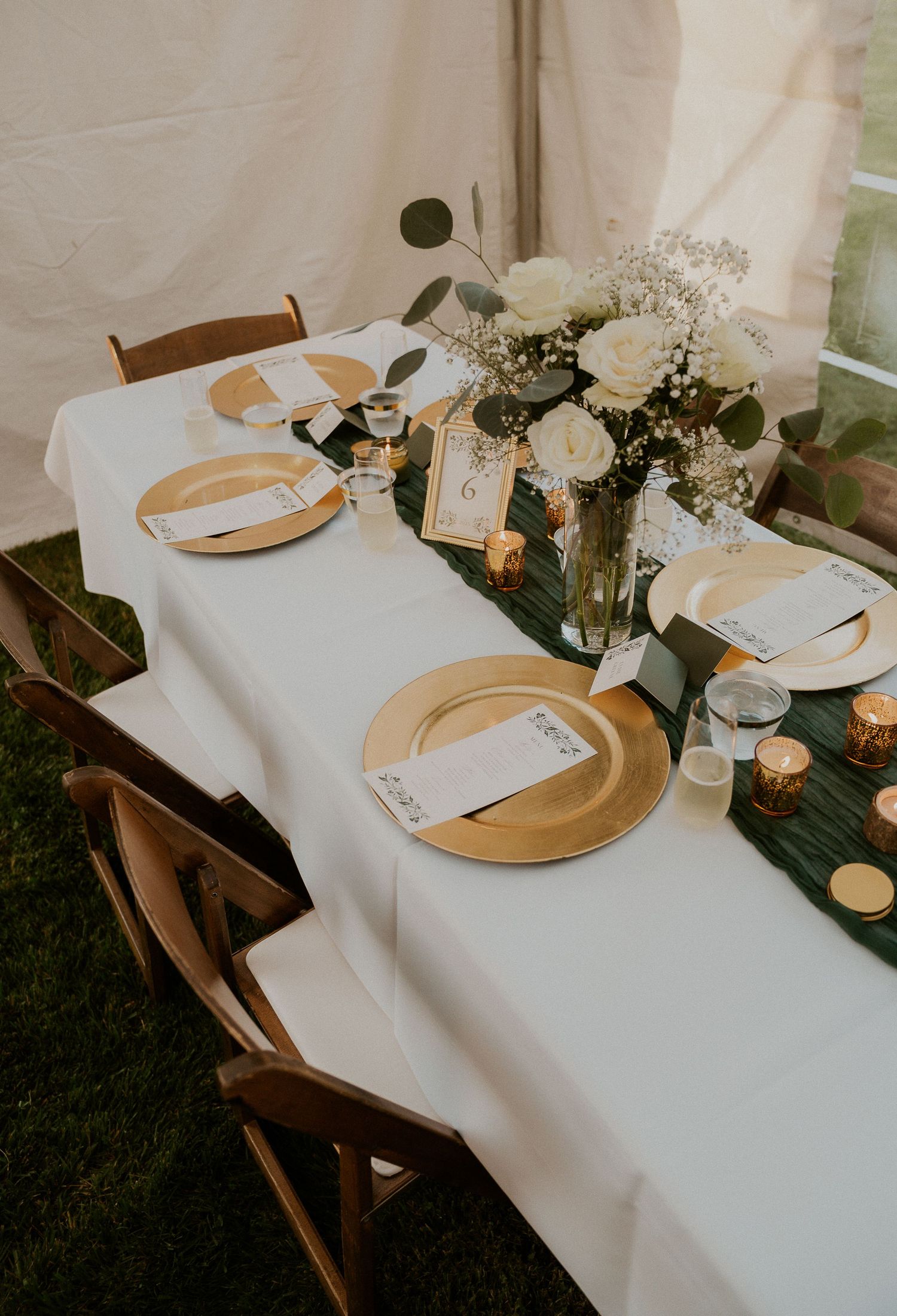 Wedding table setting with gold chargers, white linens, and elegant greenery centerpieces.