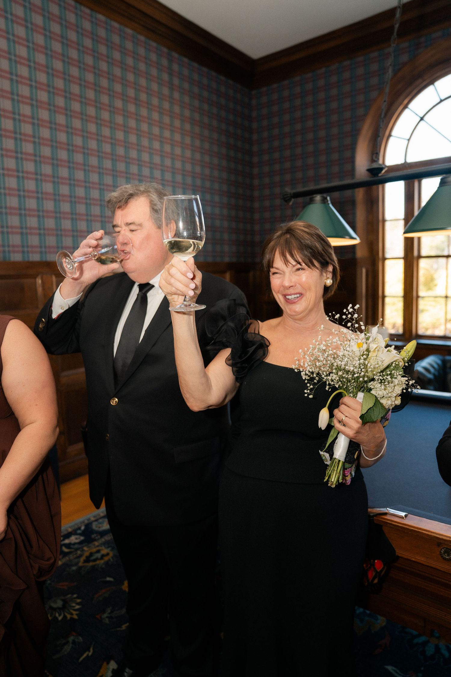 Couple in formal attire toasting with champagne, woman holding white bouquet in wood-paneled room.