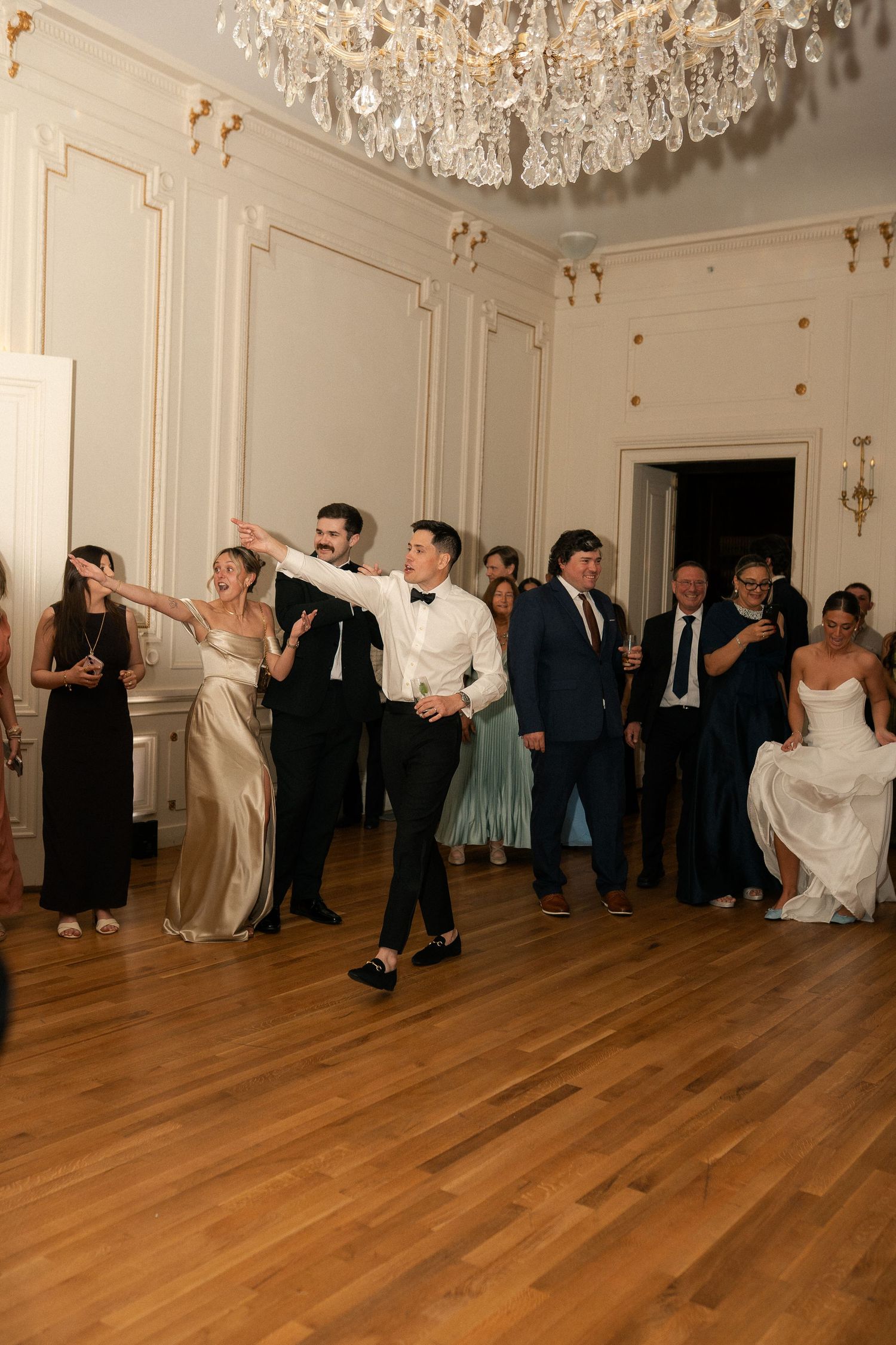 Groom dancing with wedding guests under a crystal chandelier in a grand ballroom during wedding reception.