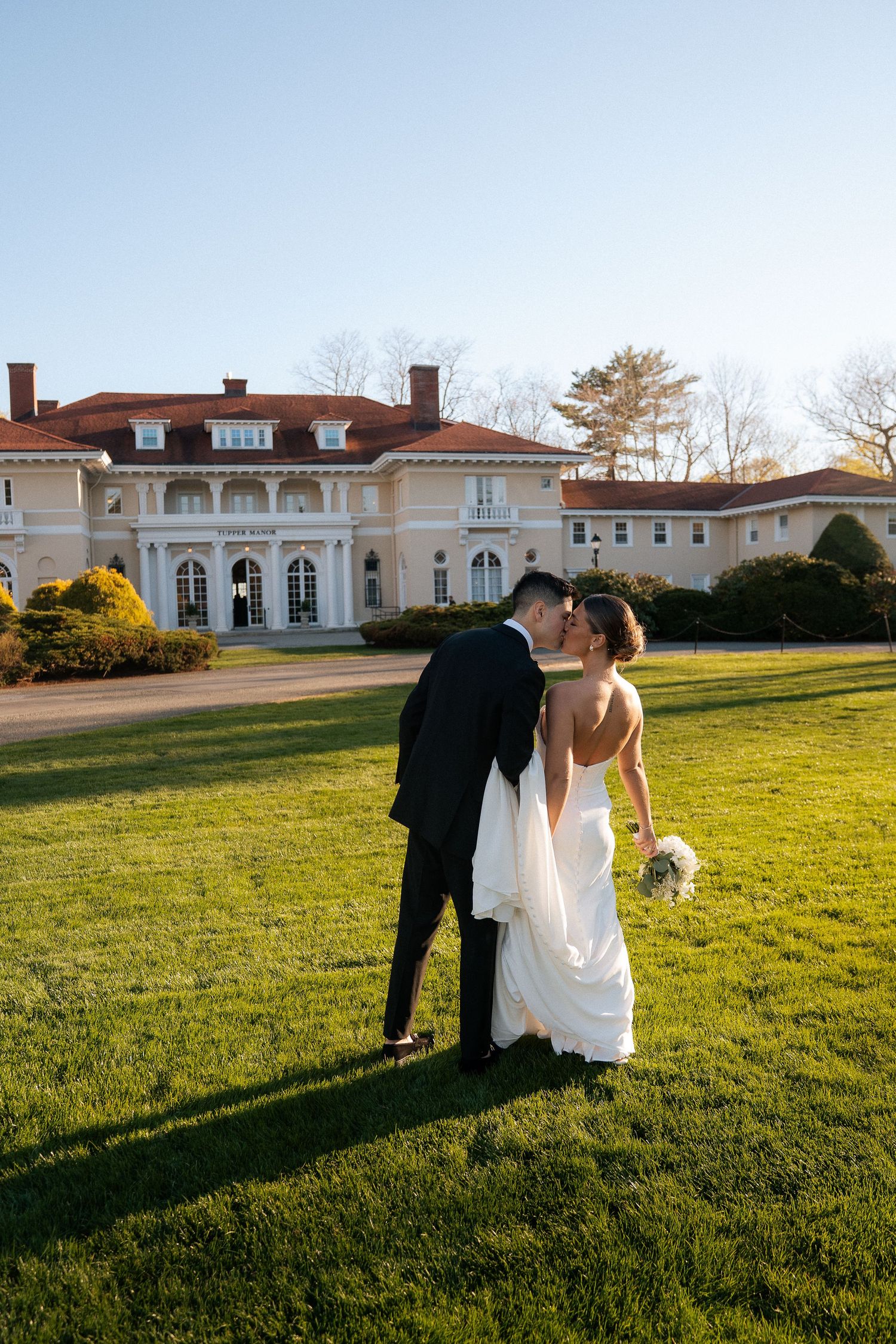 Newlyweds share a romantic kiss on the green lawn in front of a grand estate wedding venue.