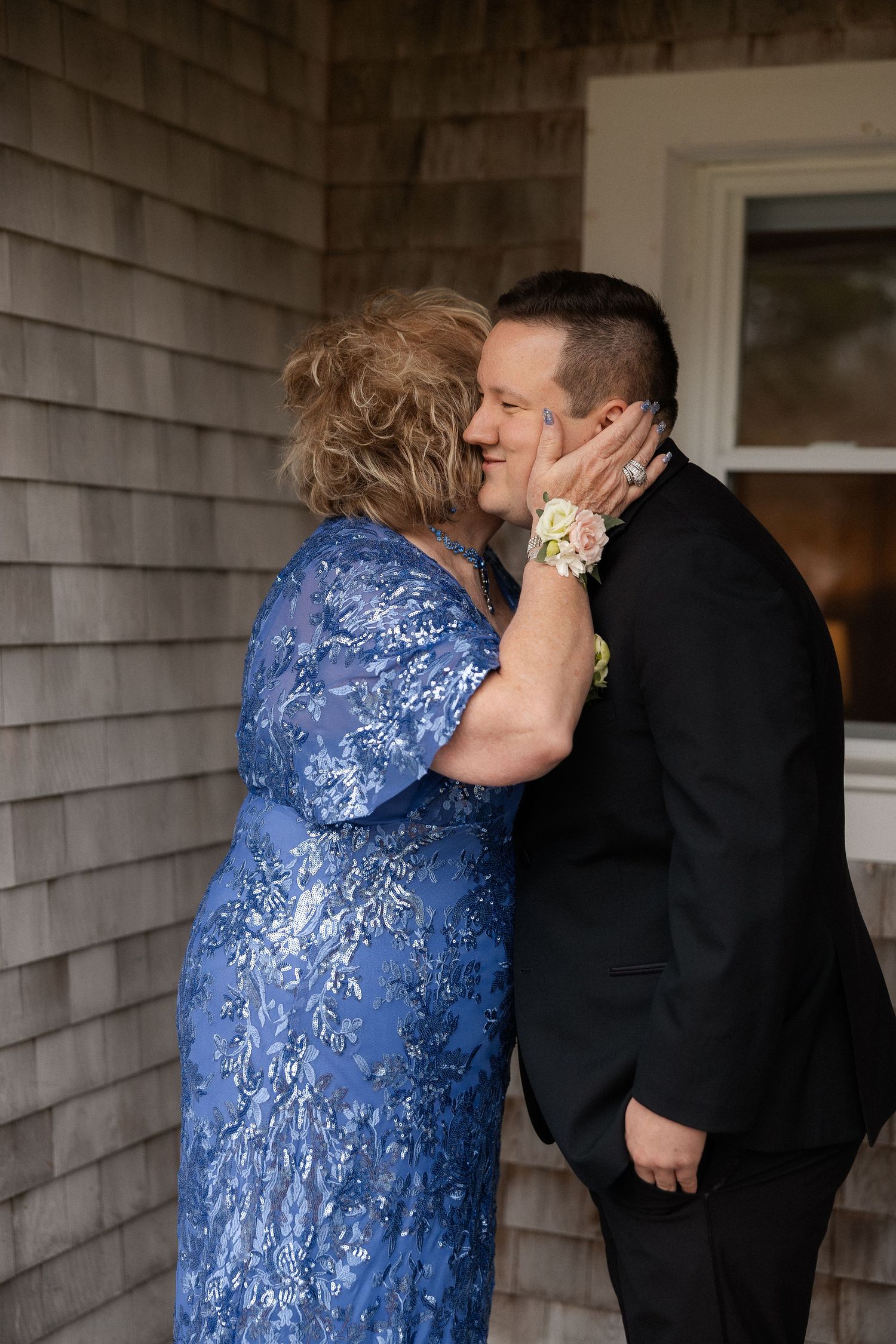 Mother in blue floral gown tenderly holding groom's face before the wedding ceremony outside.