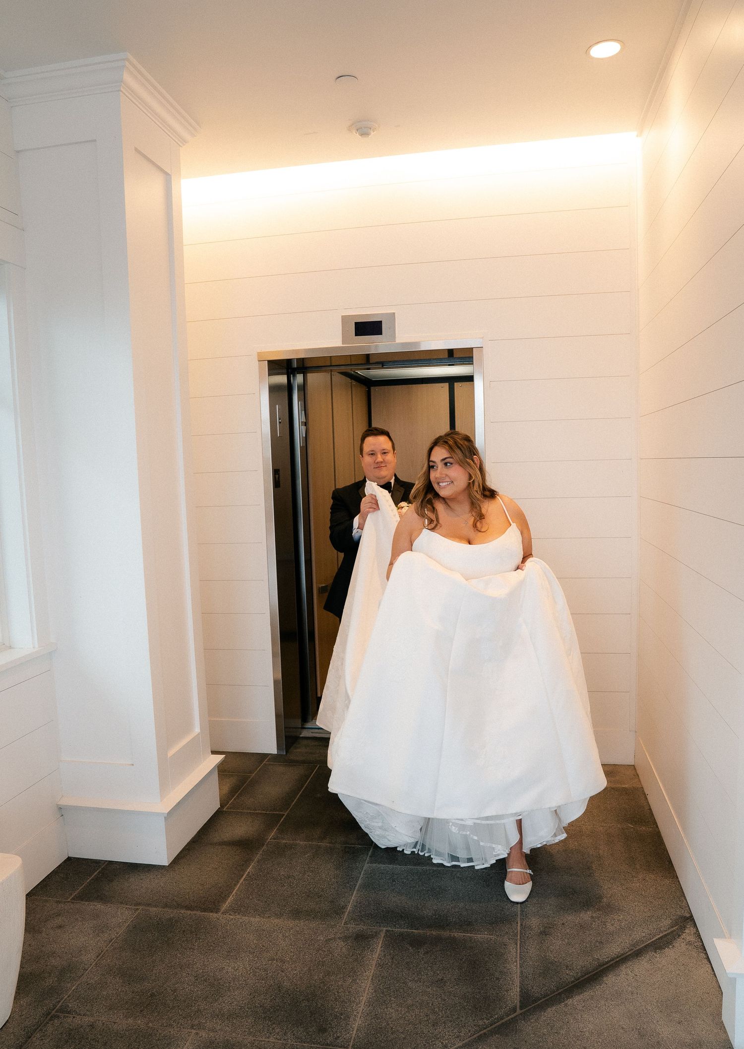 Bride and groom exit an elevator, bride holding her white wedding gown train in a white hallway.