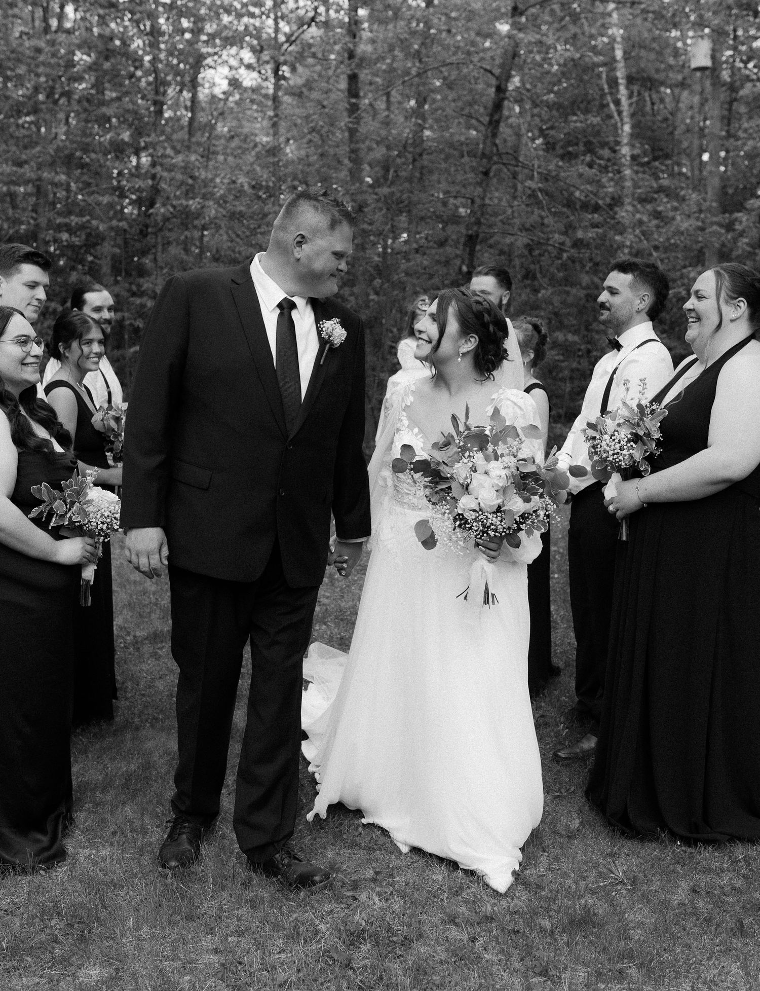 Black and white outdoor wedding photo with bridal party gathered in wooded setting.