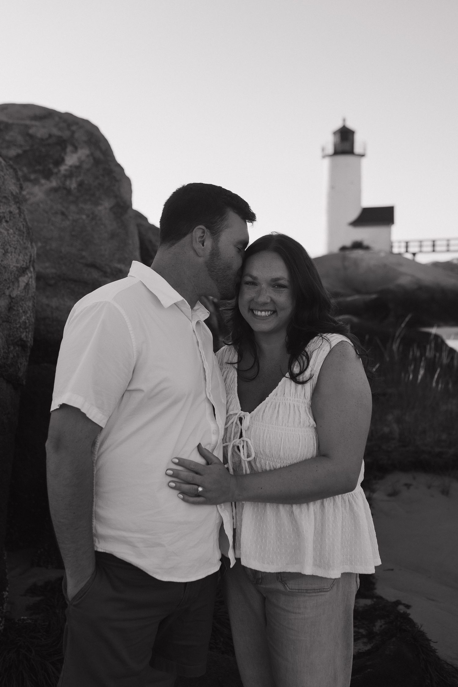 A romantic black and white photo of a couple embracing near a lighthouse on a rocky coast.