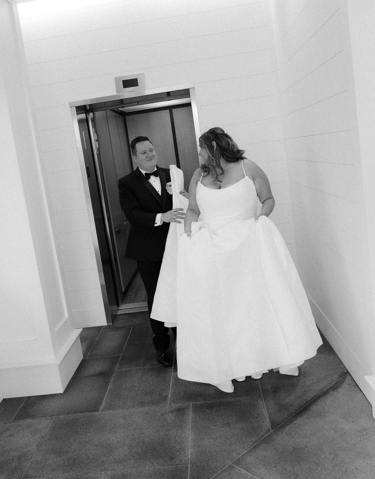 Bride and groom in formal wedding attire share a moment near an elevator in black and white photo.
