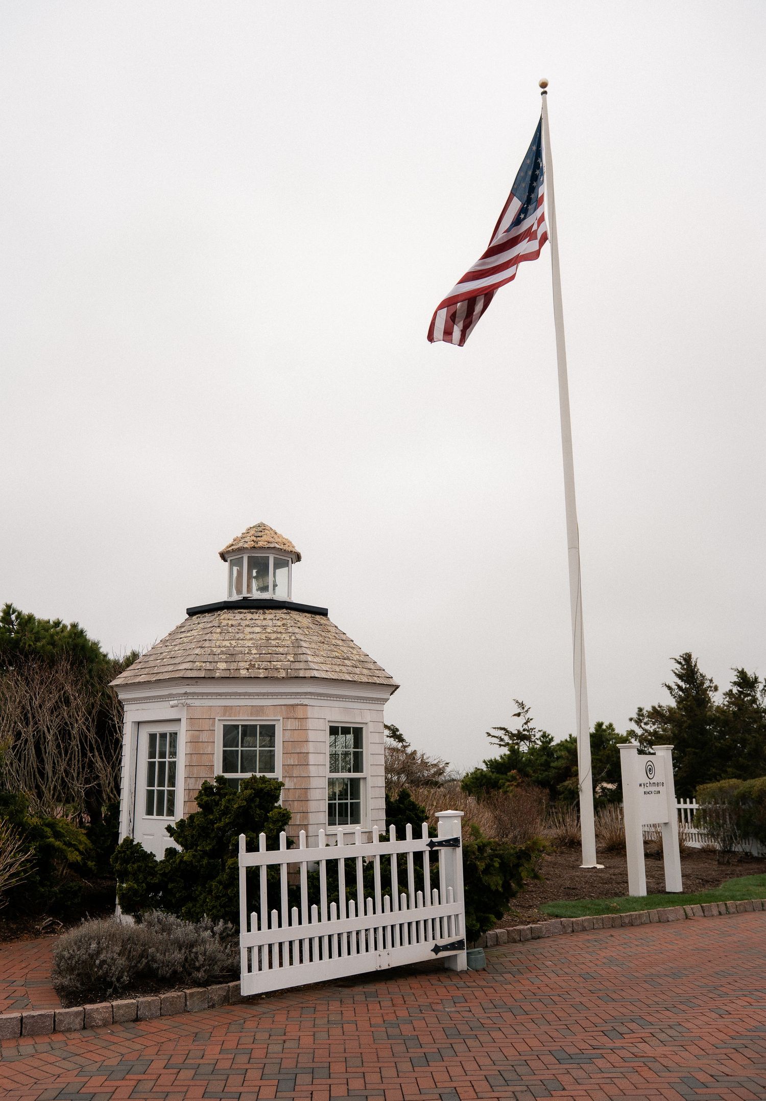 Octagonal gazebo with cedar shingles beside a tall flagpole flying the American flag on a brick path.