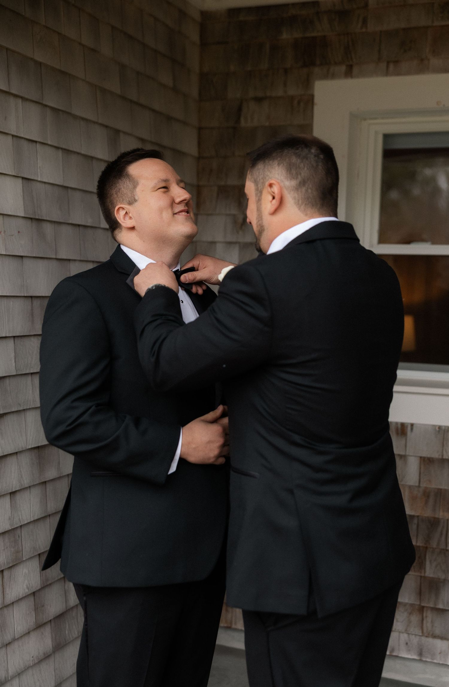 Groom smiling as groomsman adjusts his bow tie outside a shingle-style building before the wedding ceremony.