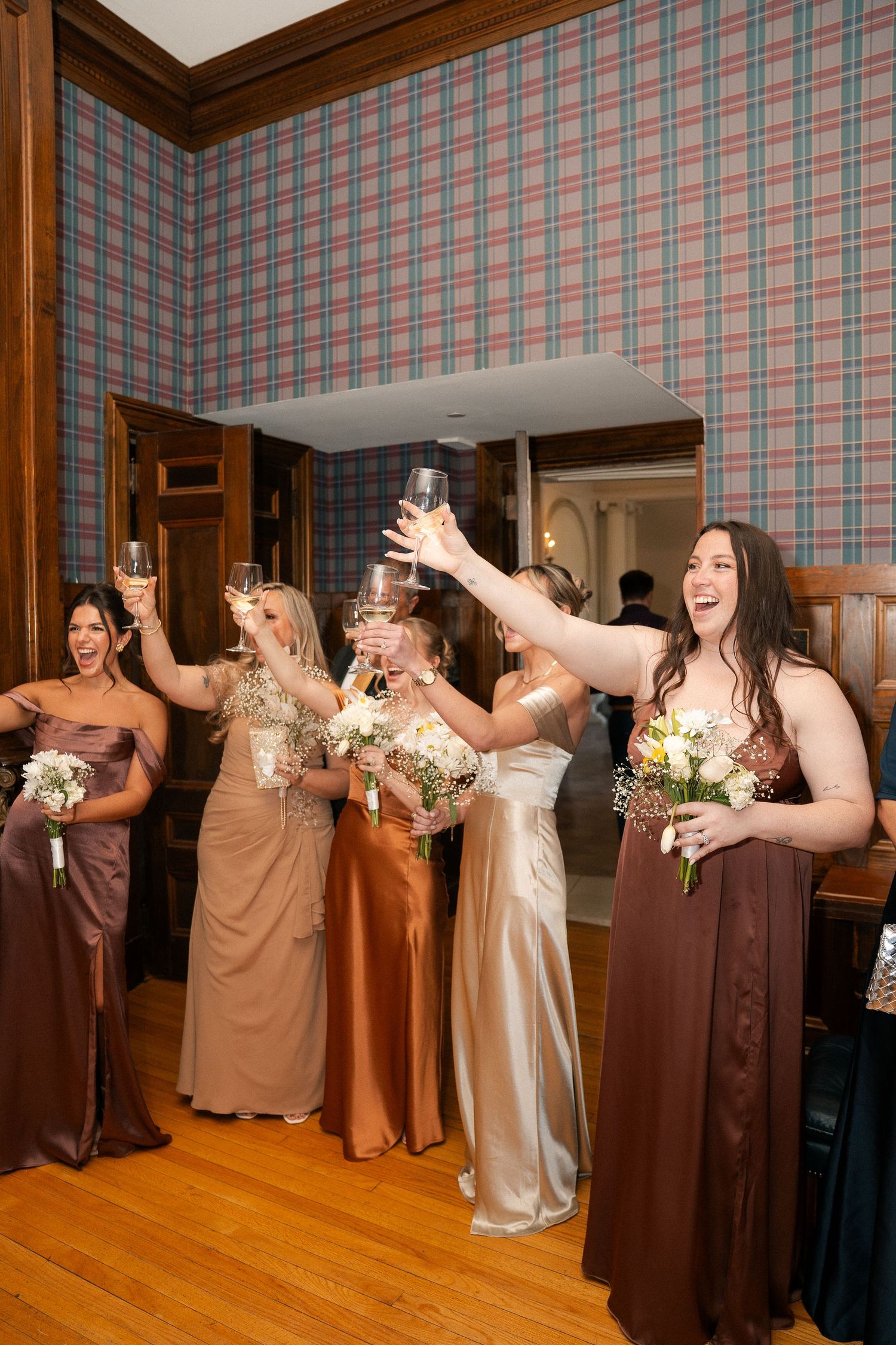 Bridesmaids in earth-tone gowns raise champagne glasses in a toast, holding floral bouquets inside a plaid-wallpapered venue.