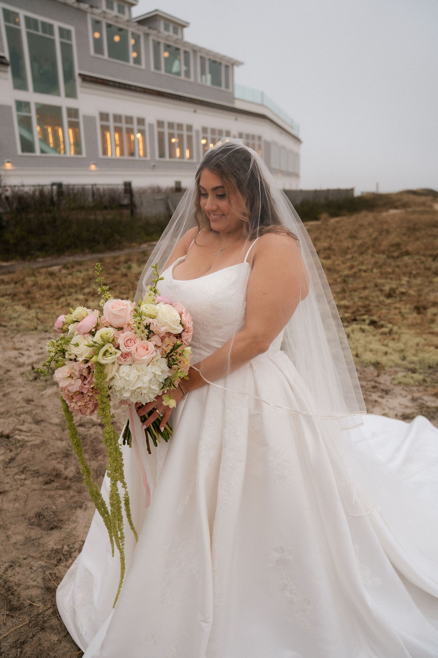 Bride in a white ball gown and veil holding a pink and white bouquet outdoors near a coastal venue.