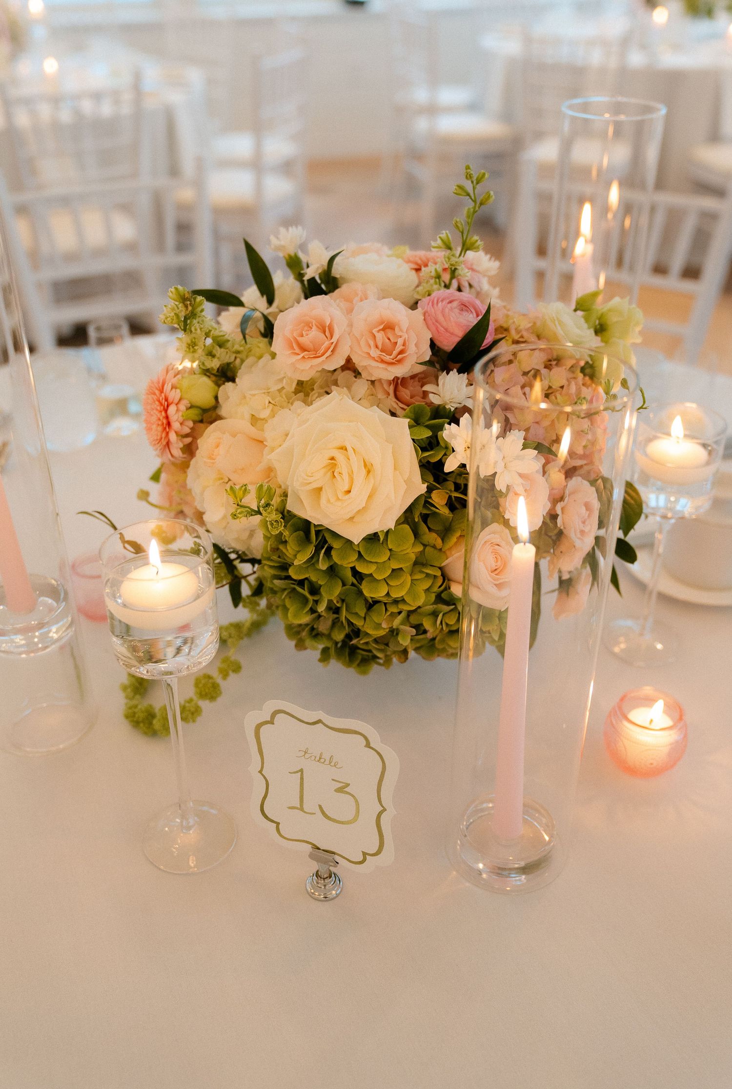 Elegant wedding centerpiece with pink roses, hydrangeas, candles, and table number 13 on a white tablecloth.