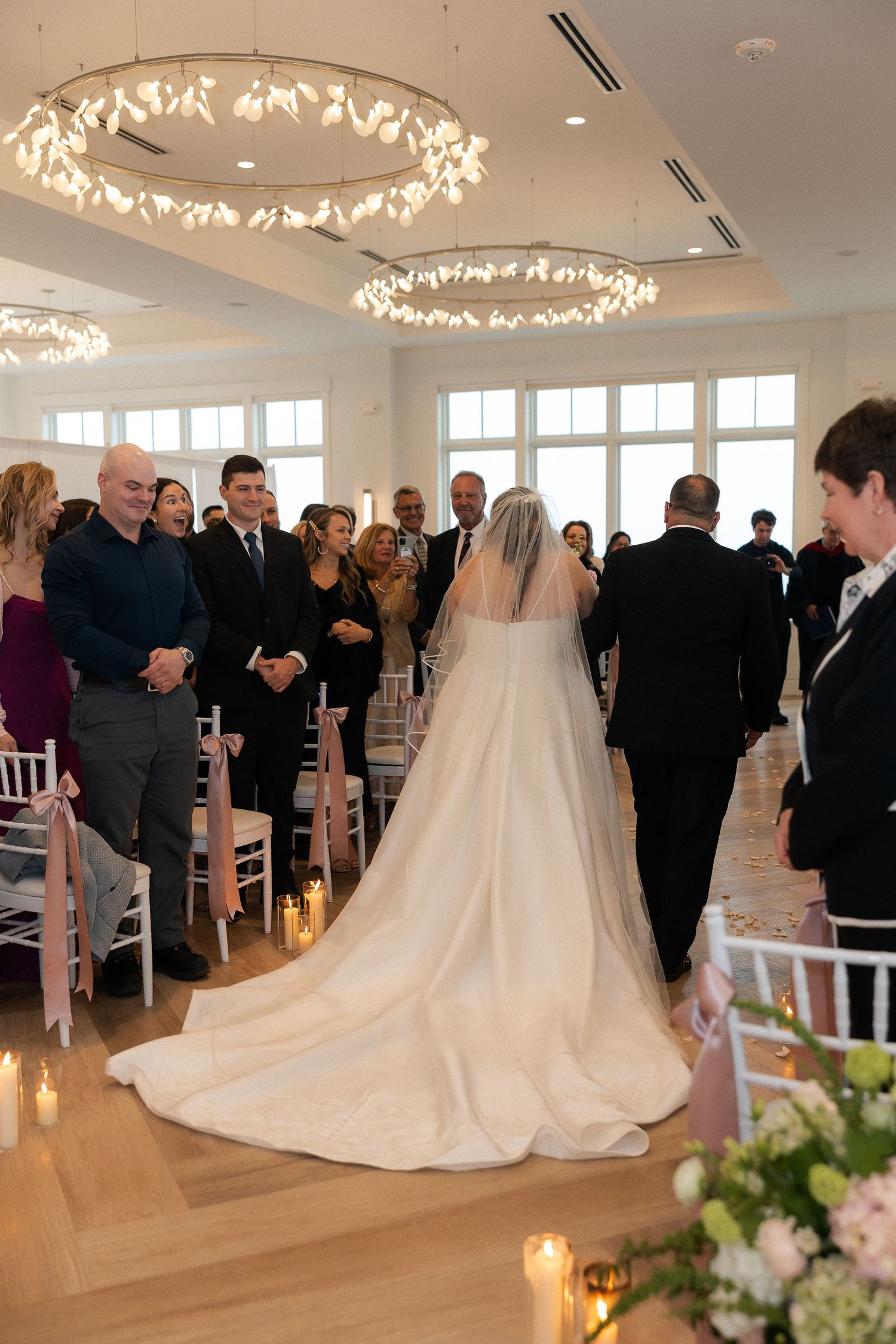 Bride in white gown with long train walks down aisle escorted by father at indoor wedding ceremony.