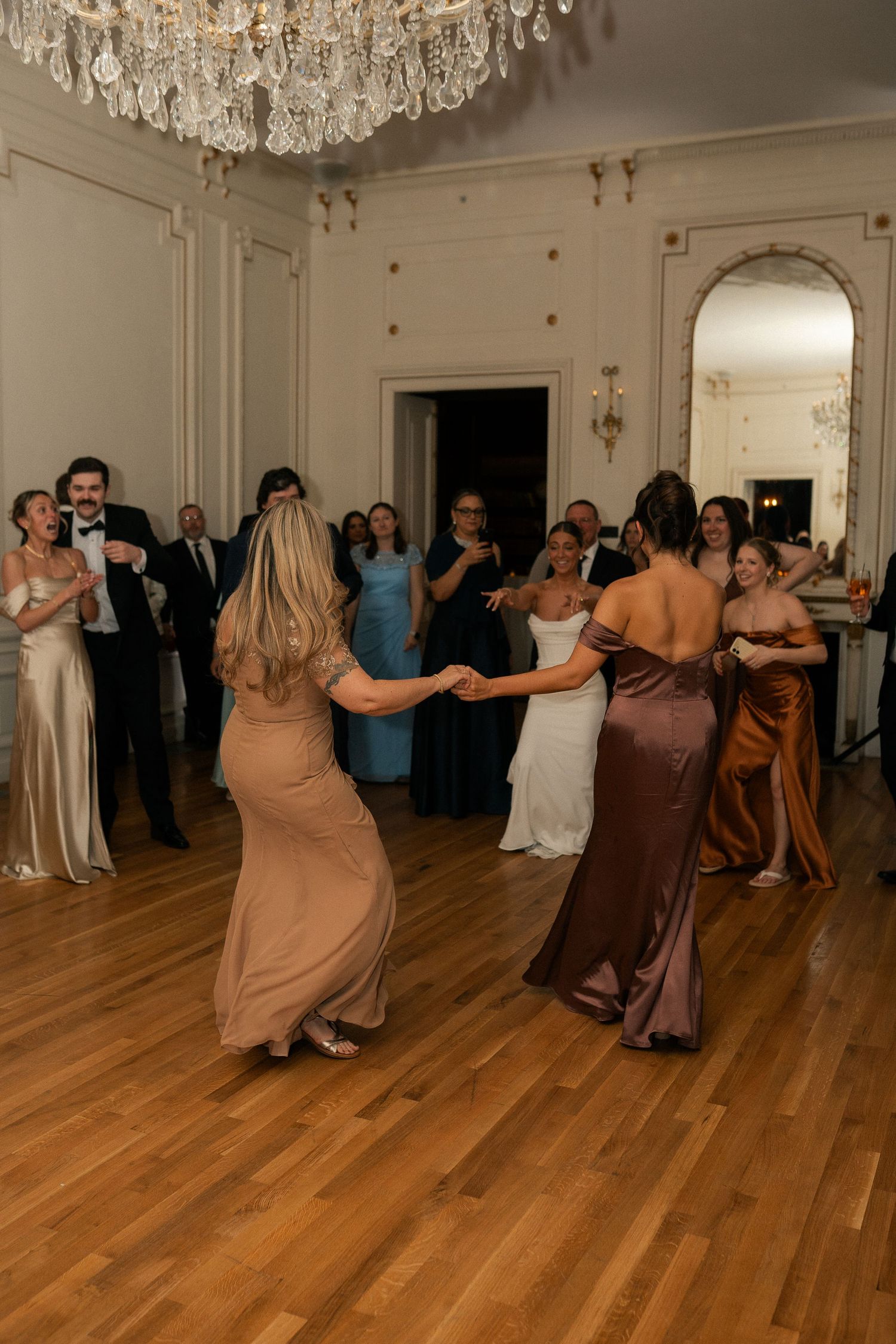 Elegant wedding reception with bridesmaids dancing in satin gowns under crystal chandelier in grand ballroom.