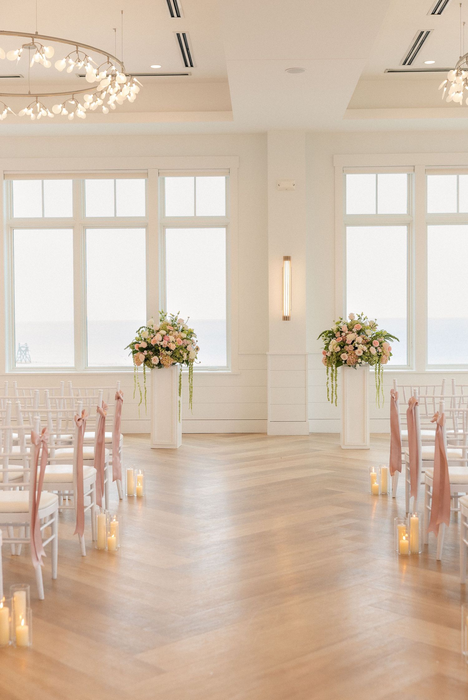 Elegant wedding ceremony setup with blush floral arrangements, white chairs, candles, and ocean views through large windows.