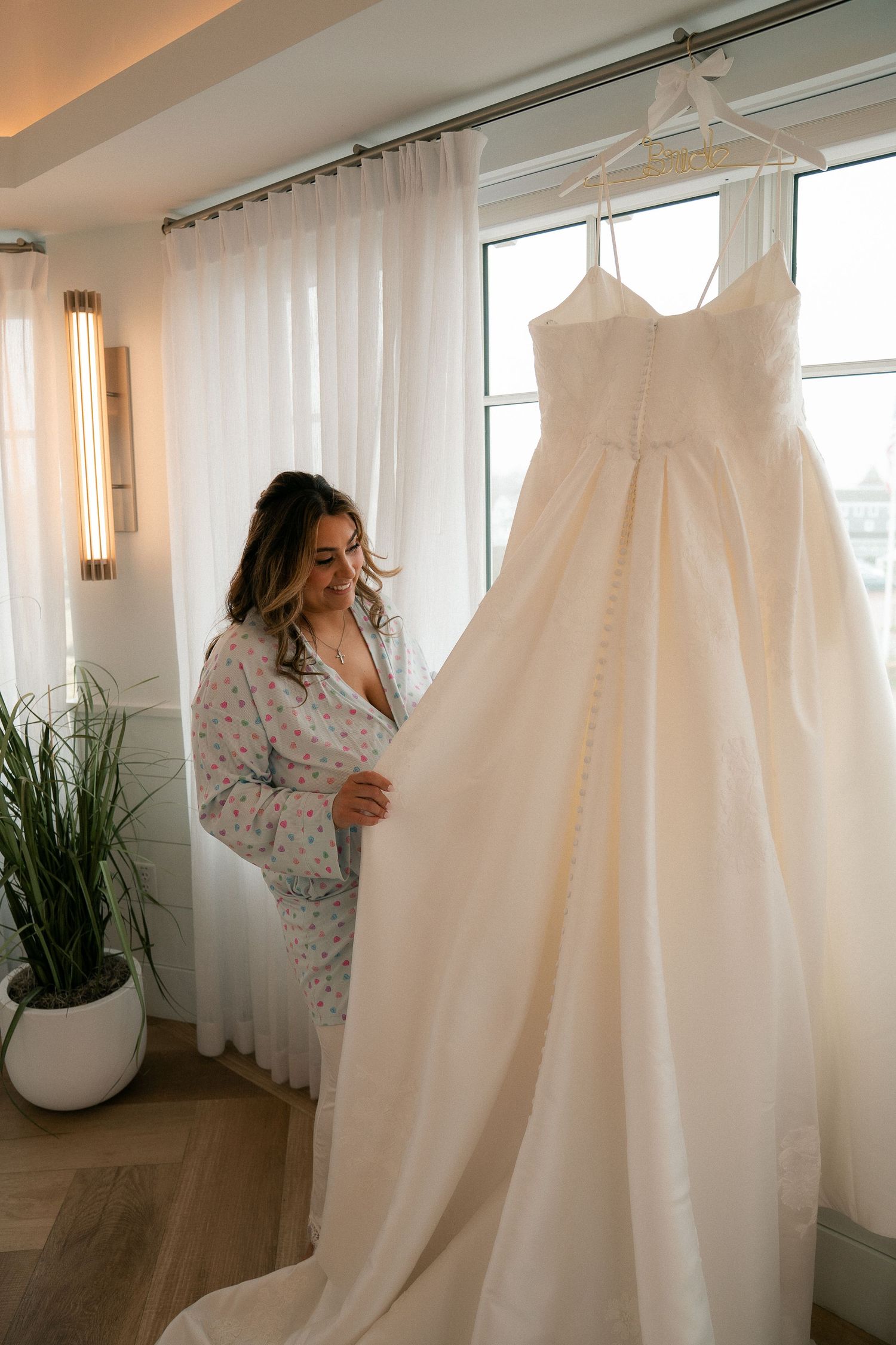 Bride in floral pajamas admiring her elegant white wedding gown hanging by a bright window in a hotel room.
