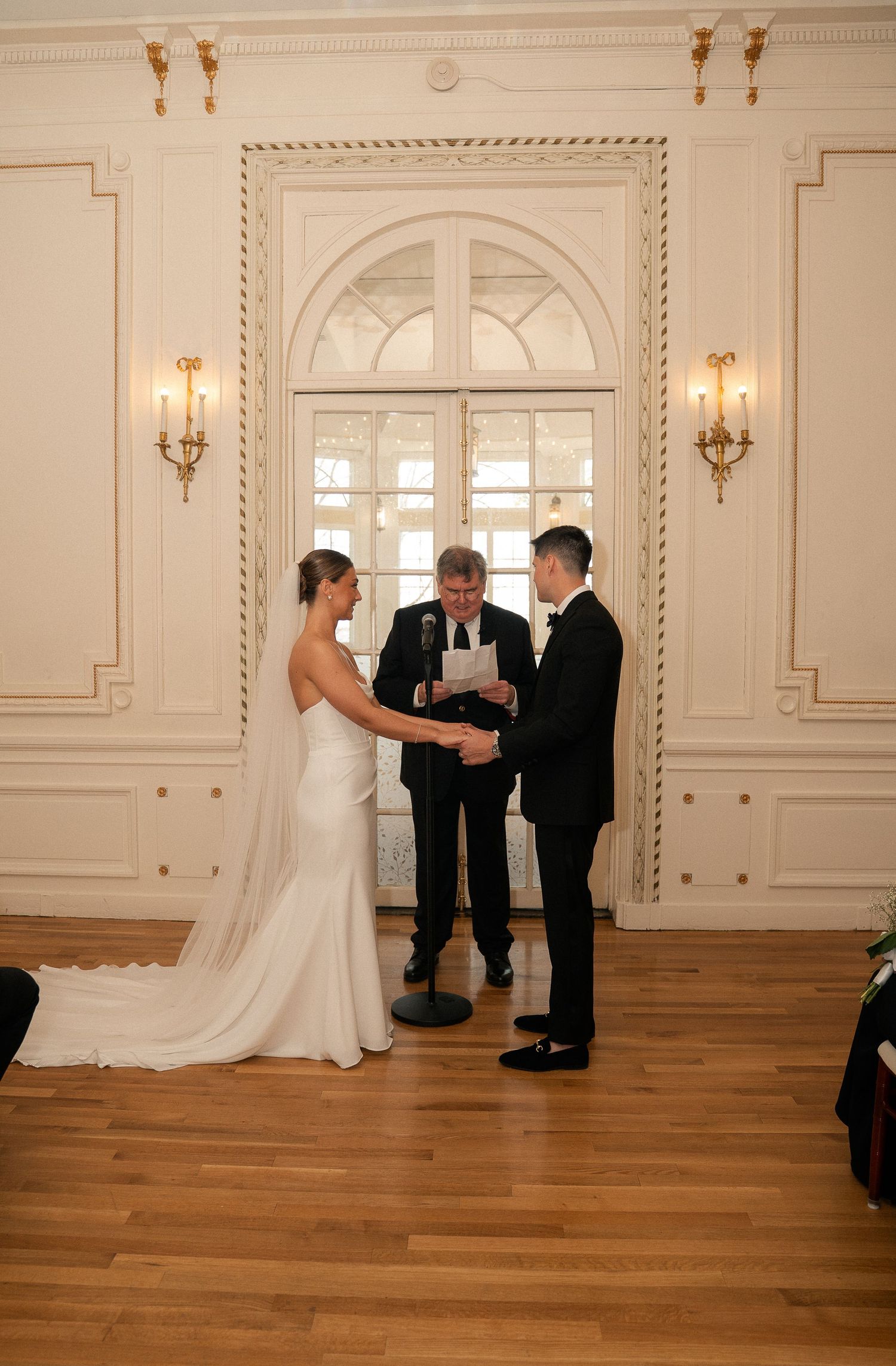 Bride and groom exchange vows before officiant in elegant white ballroom with gold sconces and arched window.
