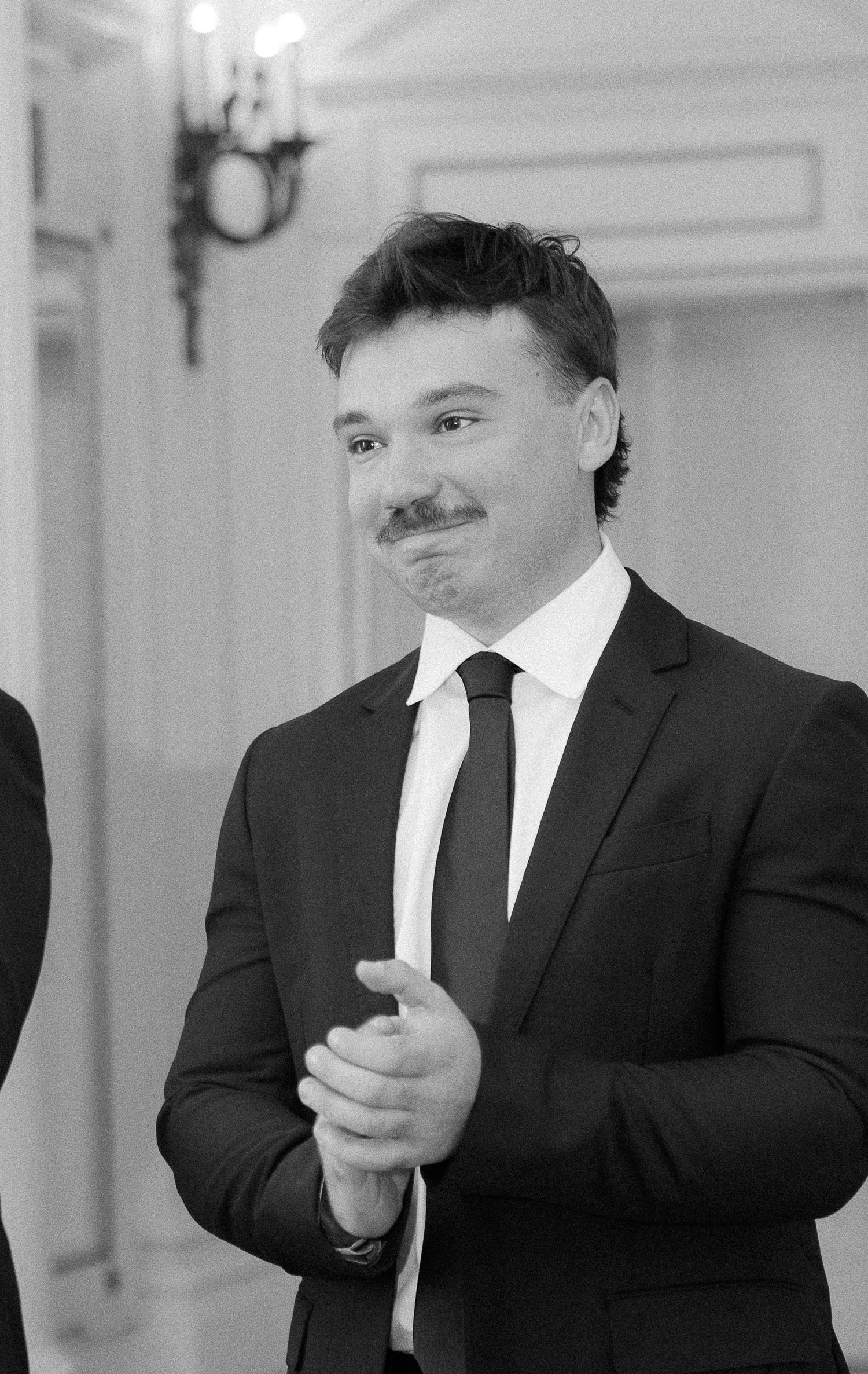 Black and white photo of a smiling man with mustache wearing a dark suit and tie, clapping hands at a formal event.
