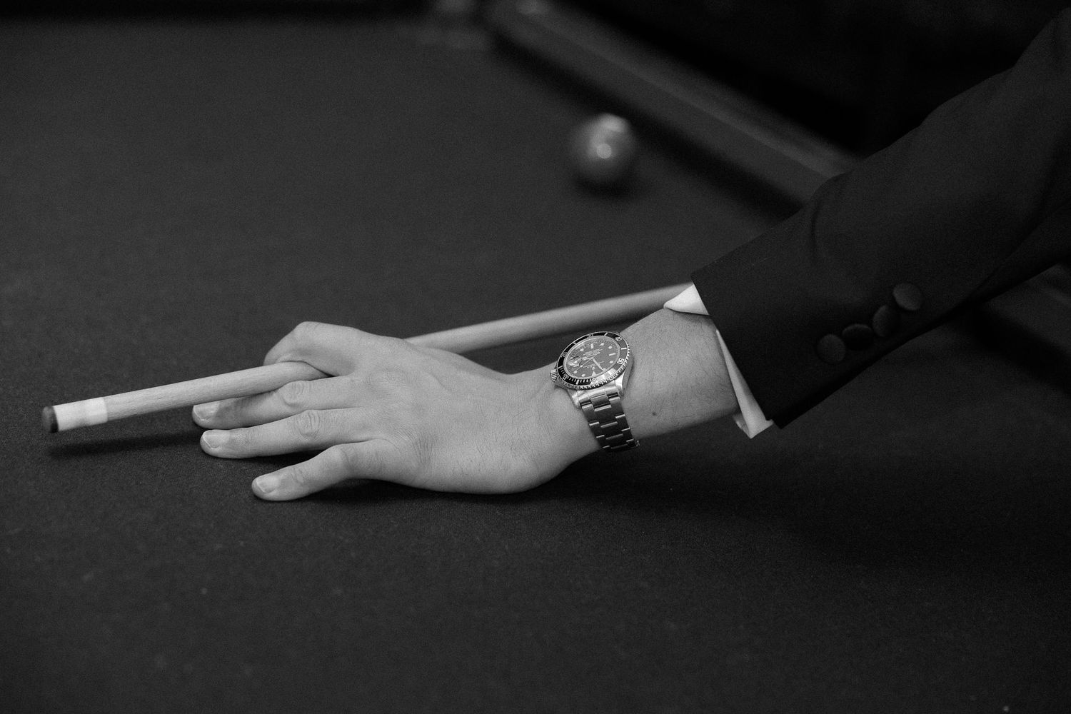 Close-up of a man in a suit wearing a luxury watch, holding a billiard cue on a pool table.